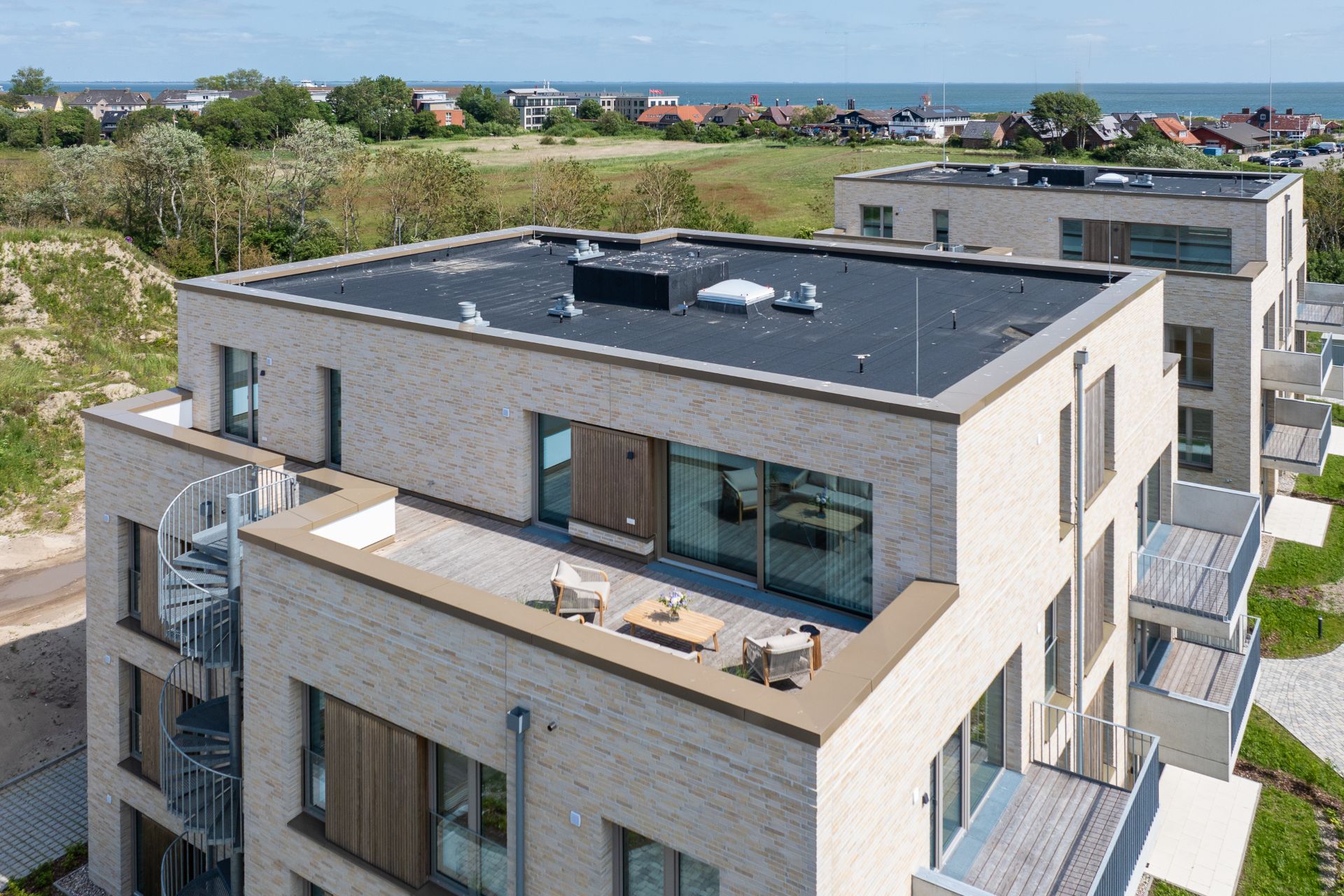 Modernes Wohngebäude mit Dachterrasse und Außentreppe, im Hintergrund grüne Landschaft und Meer.