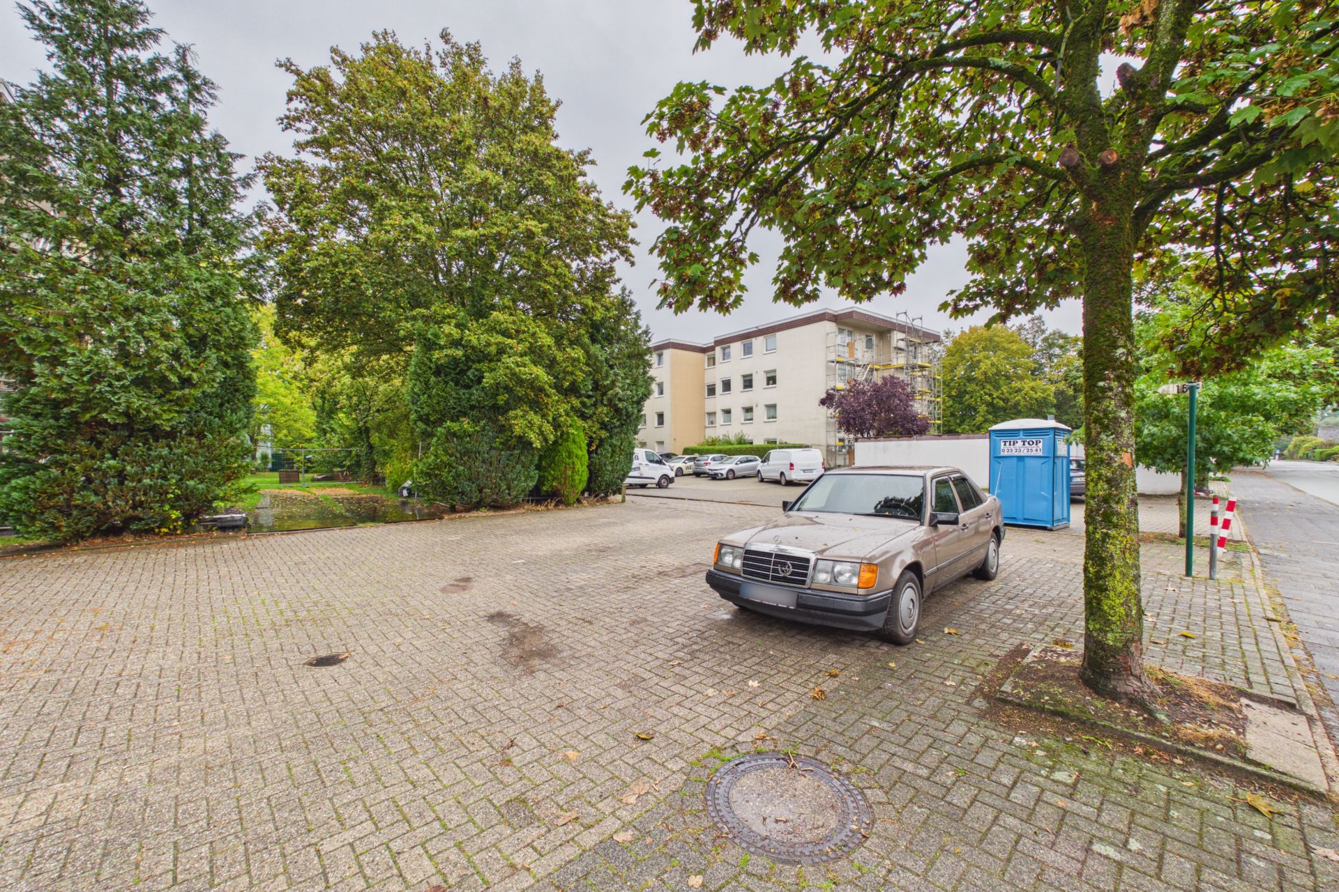 Großer gepflasterter Parkplatz mit Bäumen, geparktem Auto und Wohngebäude im Hintergrund.