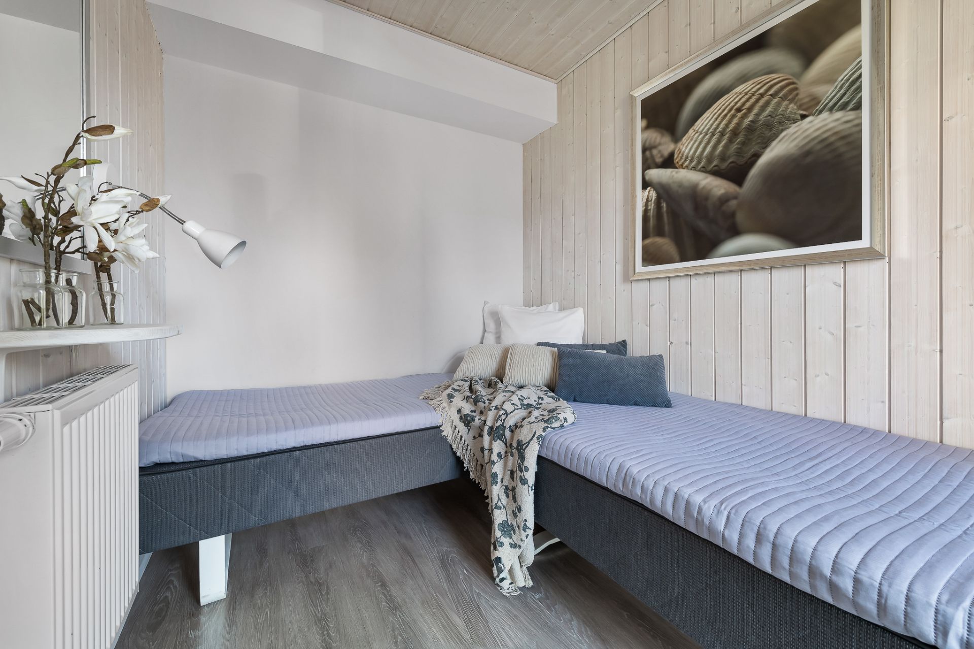 Zwei Einzelbetten in einem hellen Zimmer mit Kissen, Bild an Holzwand und Dekovase mit Blumen.
