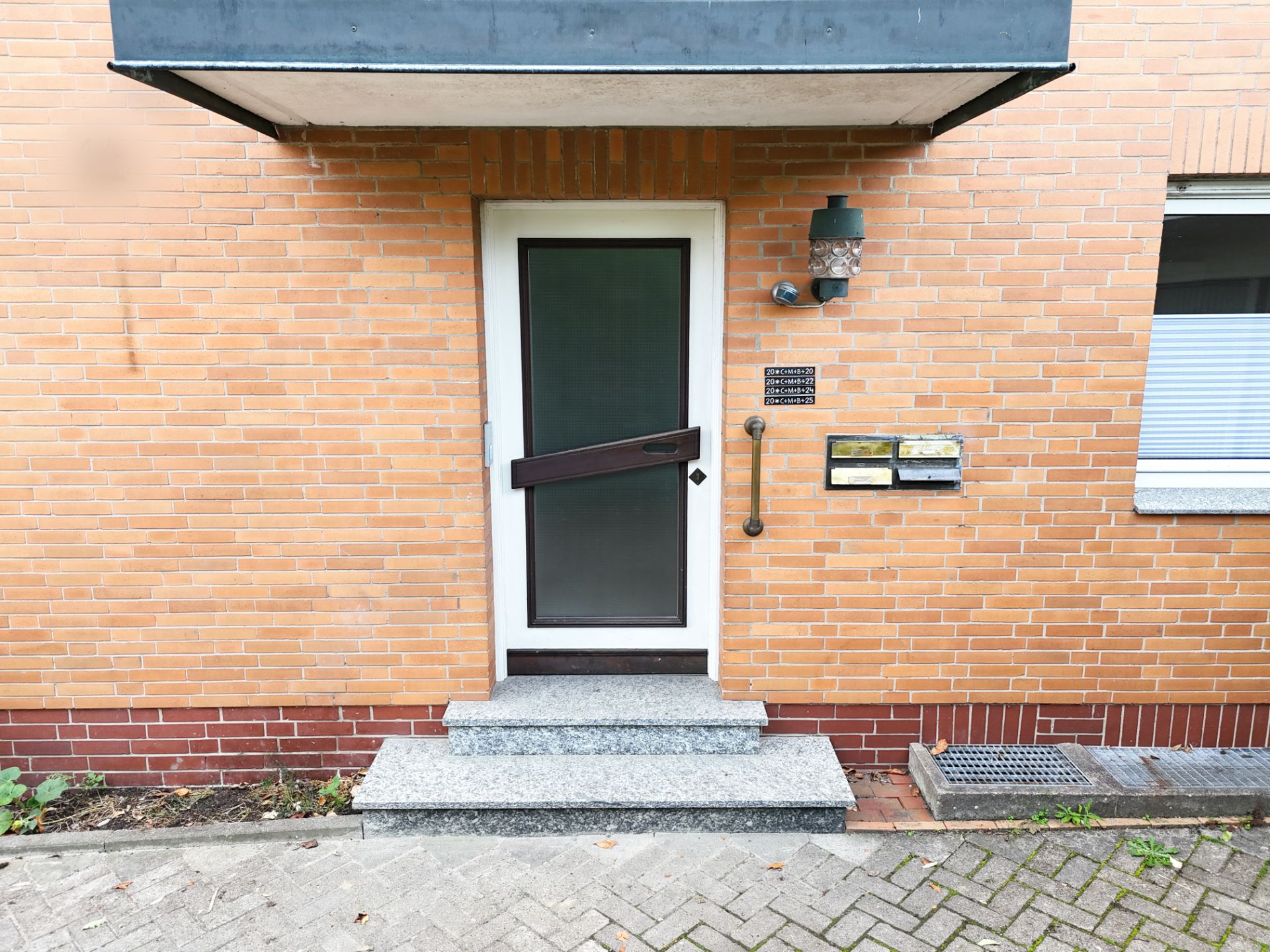 Hauseingang mit Glastür, Klingel, Briefkästen und Stufen vor einer roten Ziegelwand.