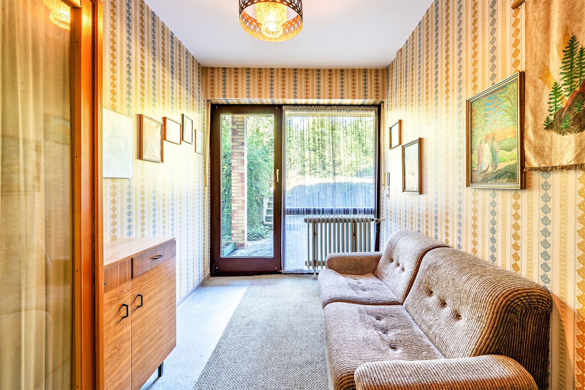 Schmaler Wohnraum mit braunem Sofa, Holzkommode, Bildern an der Wand und großem Fenster zur Terrasse.