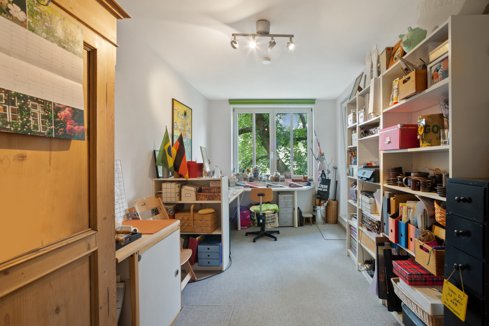 Helles Arbeitszimmer mit Schreibtisch am Fenster, Regalen und vielen Bastelmaterialien.