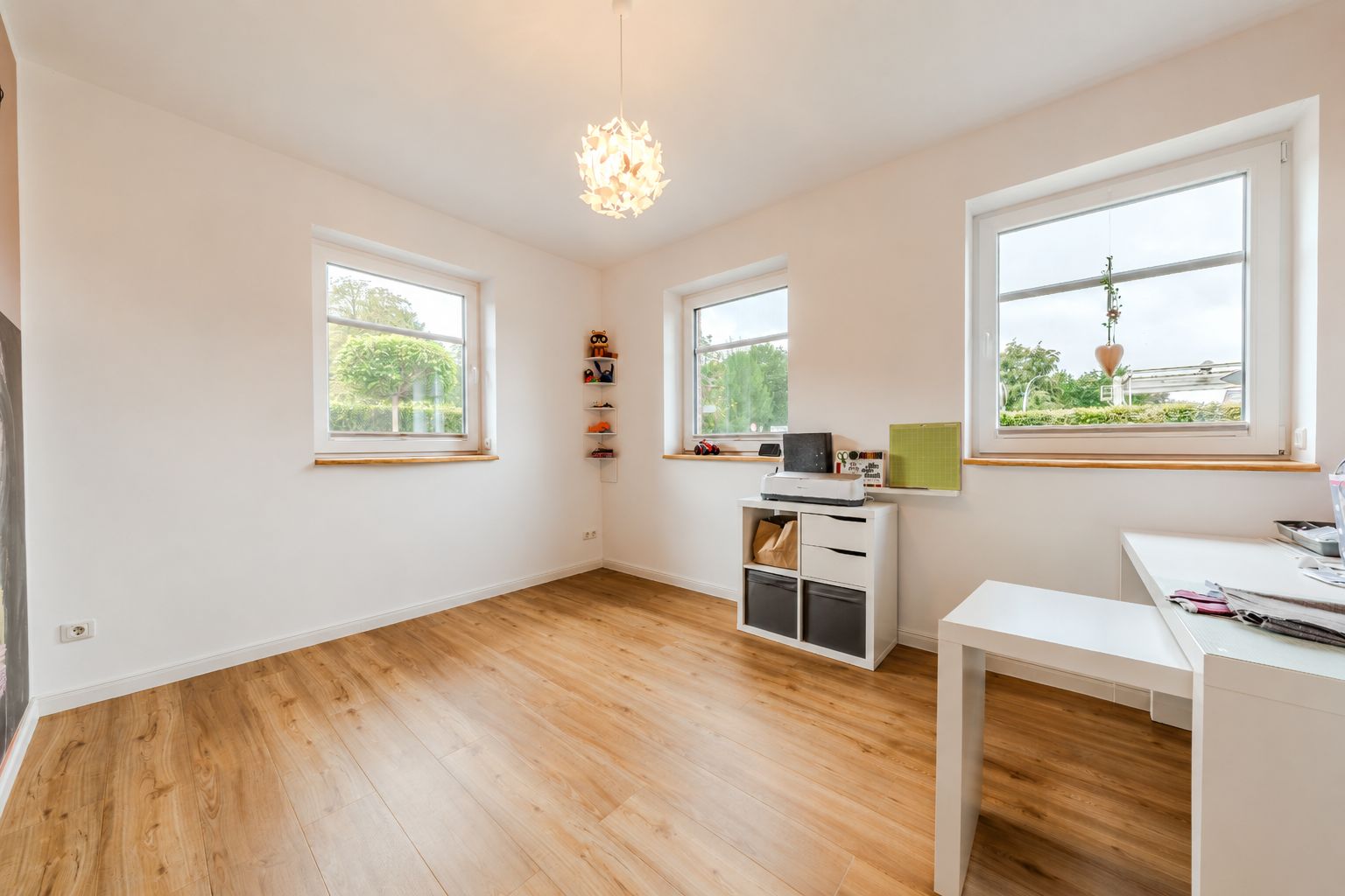 Helles Zimmer mit Holzfußboden, drei Fenstern, Schreibtisch und Regal.