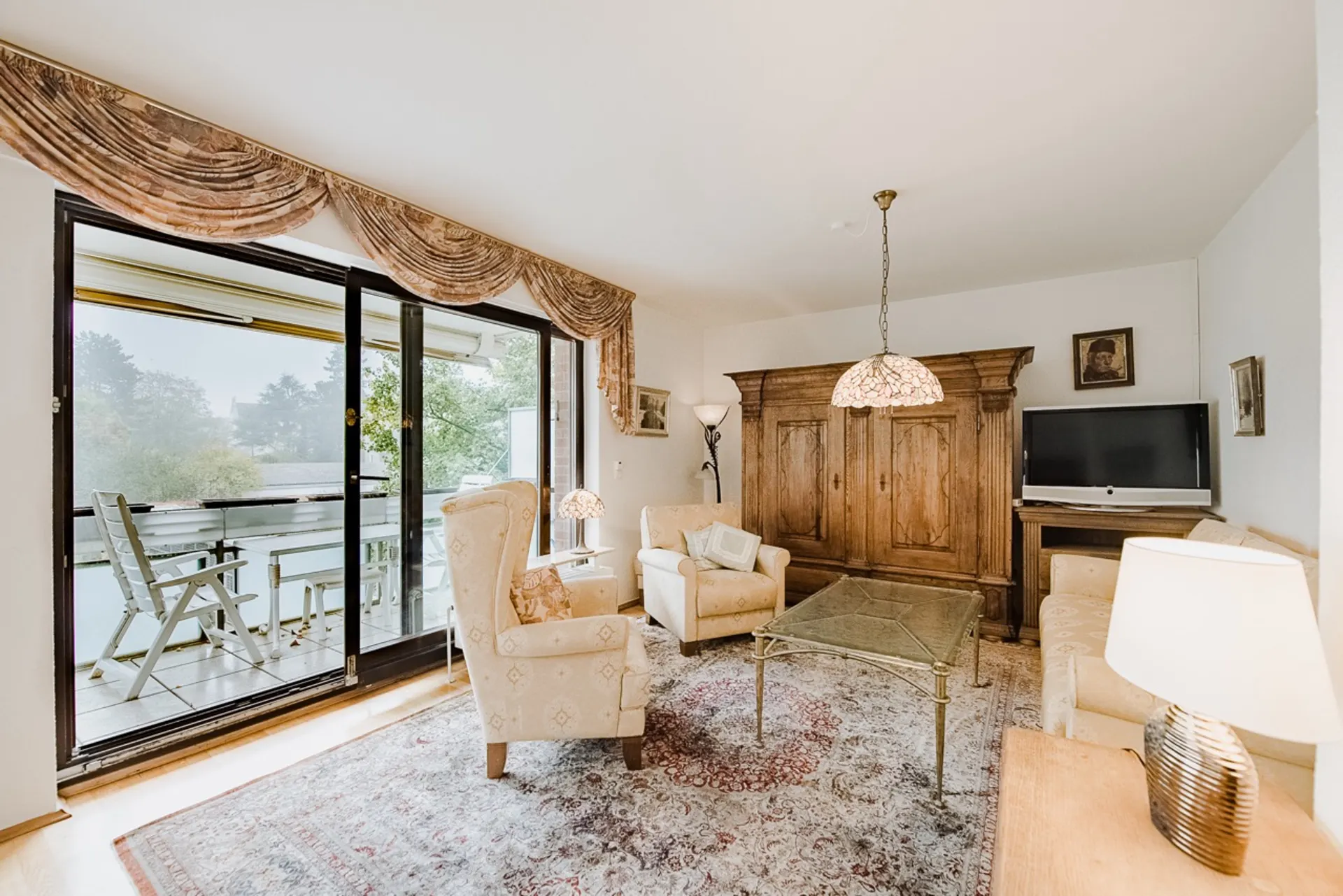 Wohnzimmer mit großen Fenstern, Sofa, Sesseln, Glastisch, Teppich und Zugang zum Balkon.