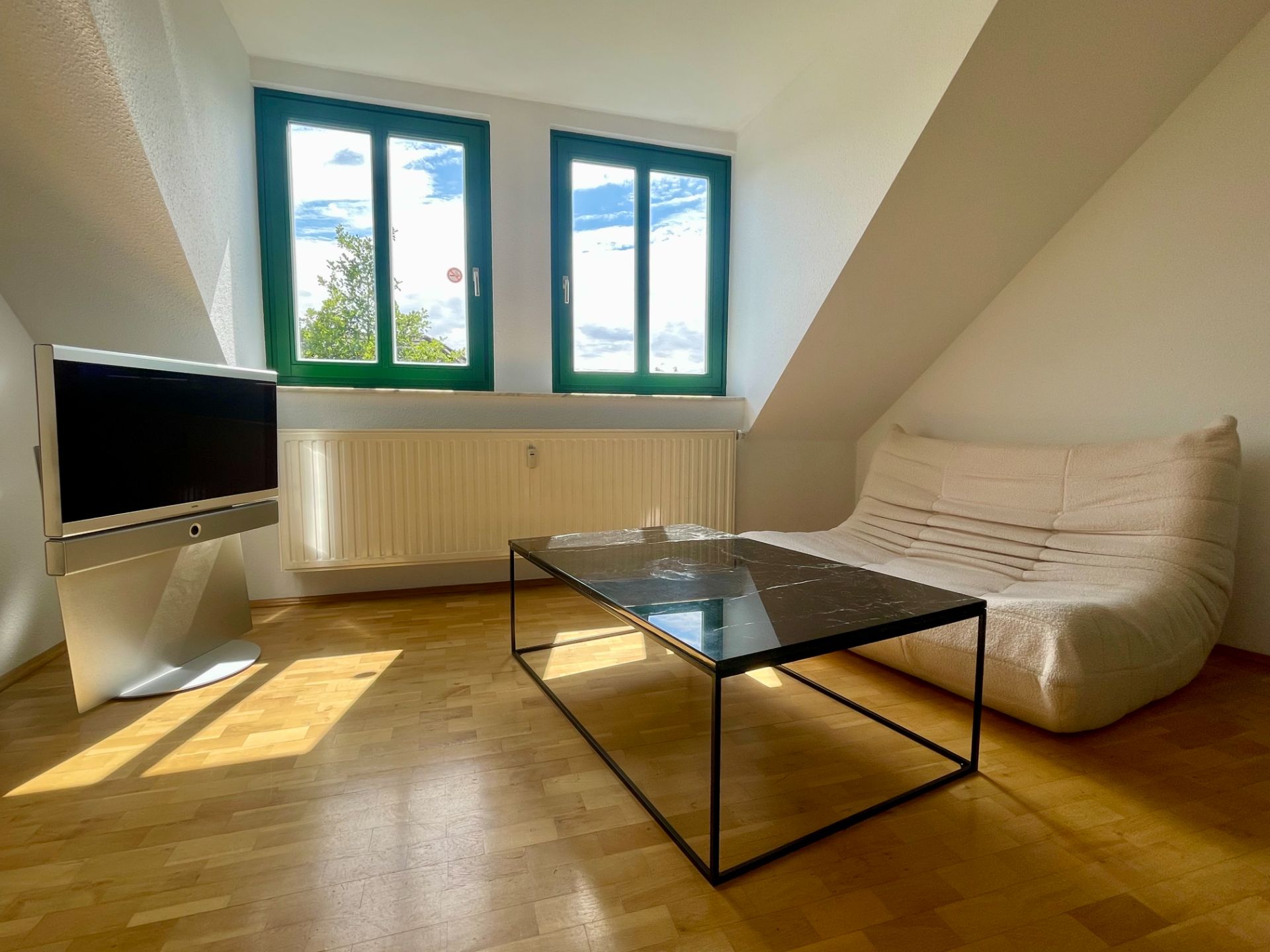 Wohnzimmer mit weißem Sofa, schwarzem Tisch, Fernseher und zwei Fenstern mit grünem Rahmen.