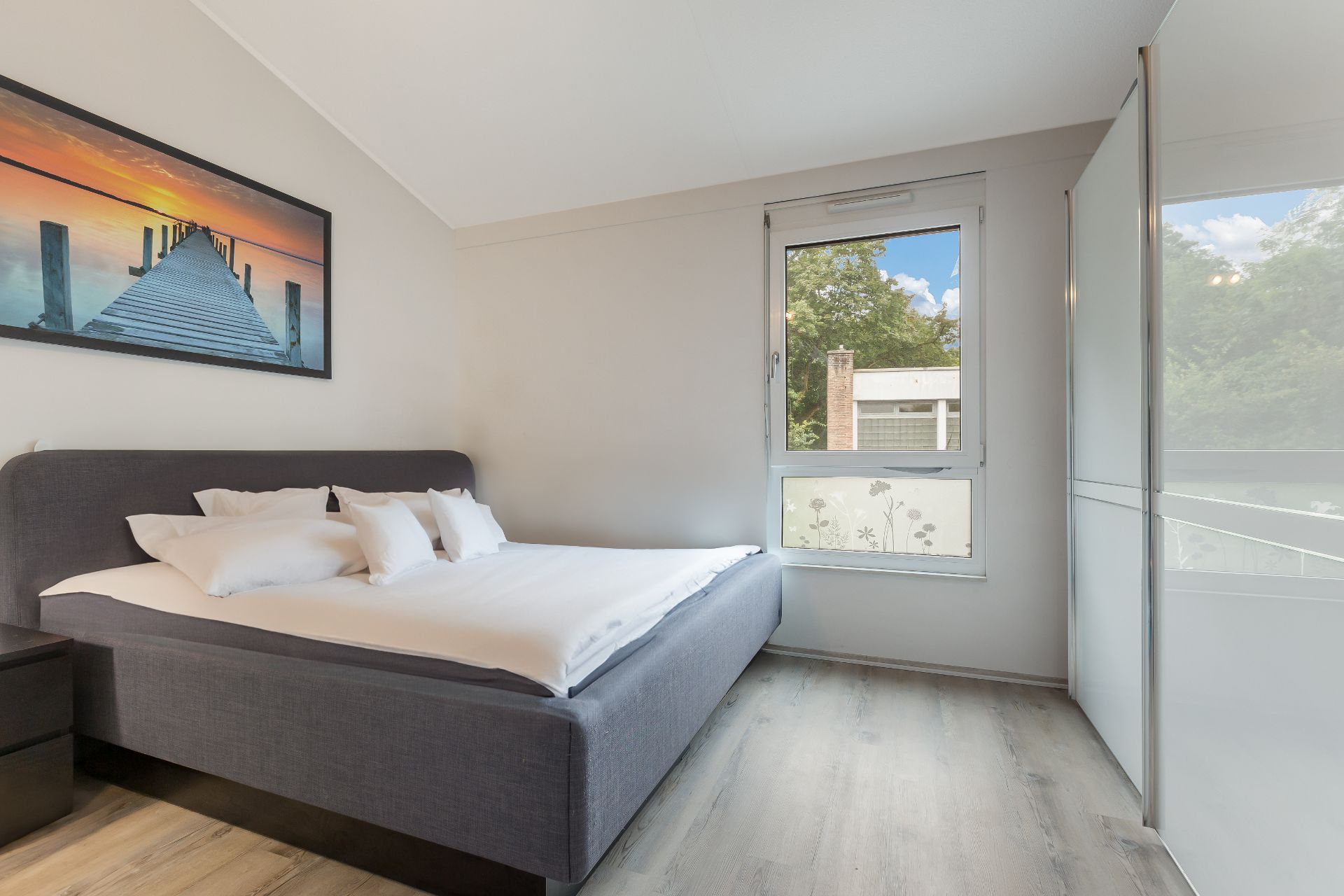 Helles Schlafzimmer mit großem Bett, Fenster, Kleiderschrank und Wandbild mit Sonnenuntergang.