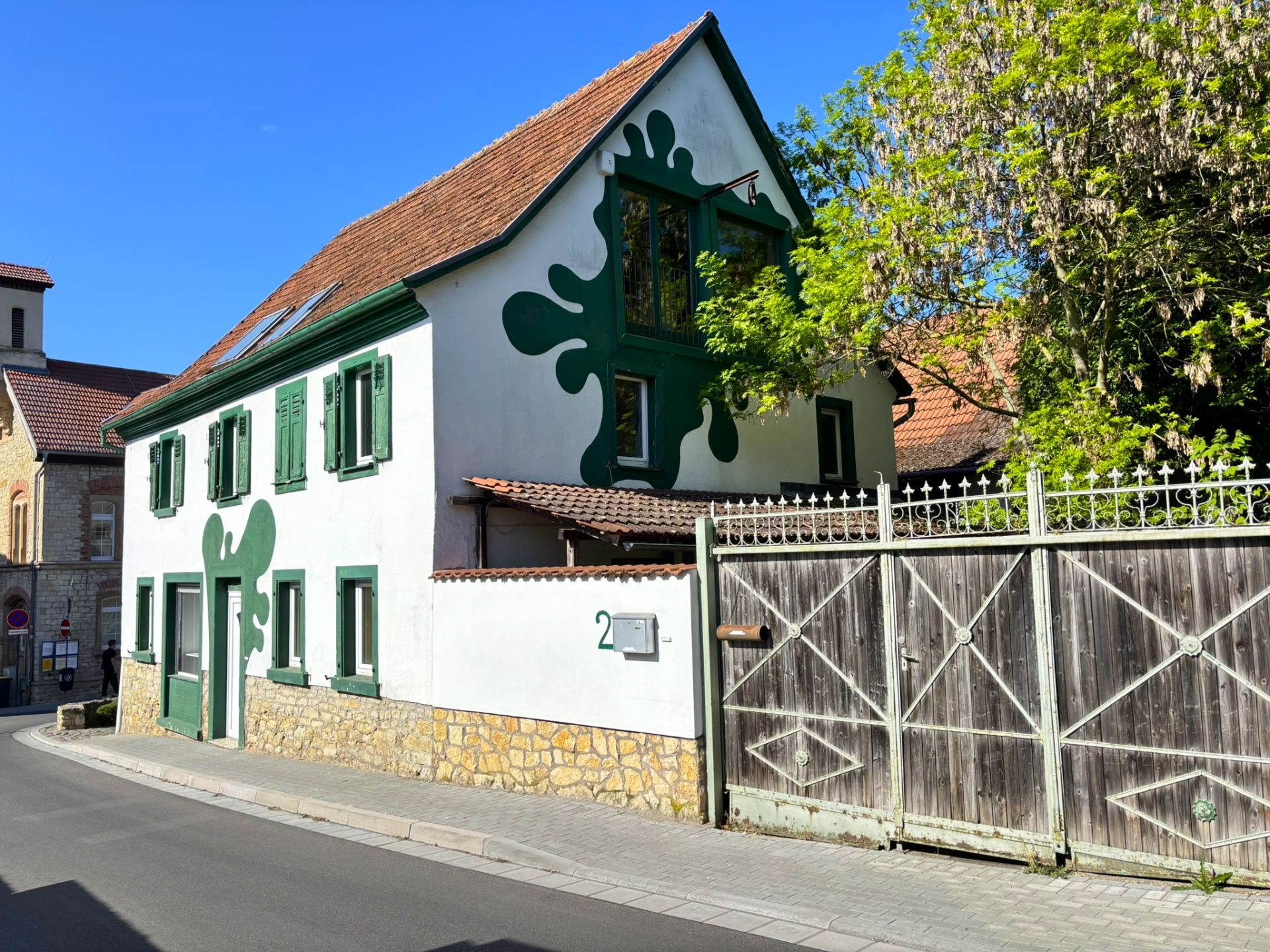 Weißes Haus mit grünen Fensterläden und abstrakten grünen Mustern an der Fassade, daneben Holztor.