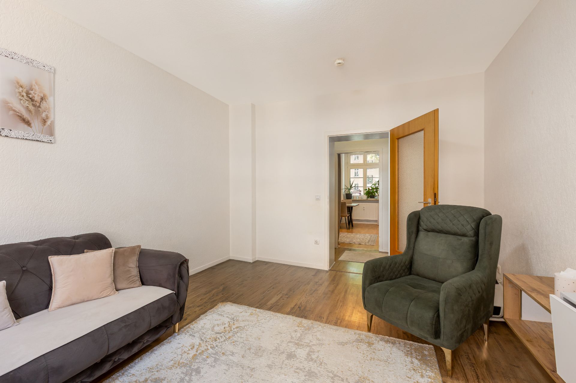 Wohnzimmer mit grauem Sofa, grünem Sessel und Teppich, Holzboden und offener Tür zum nächsten Raum.