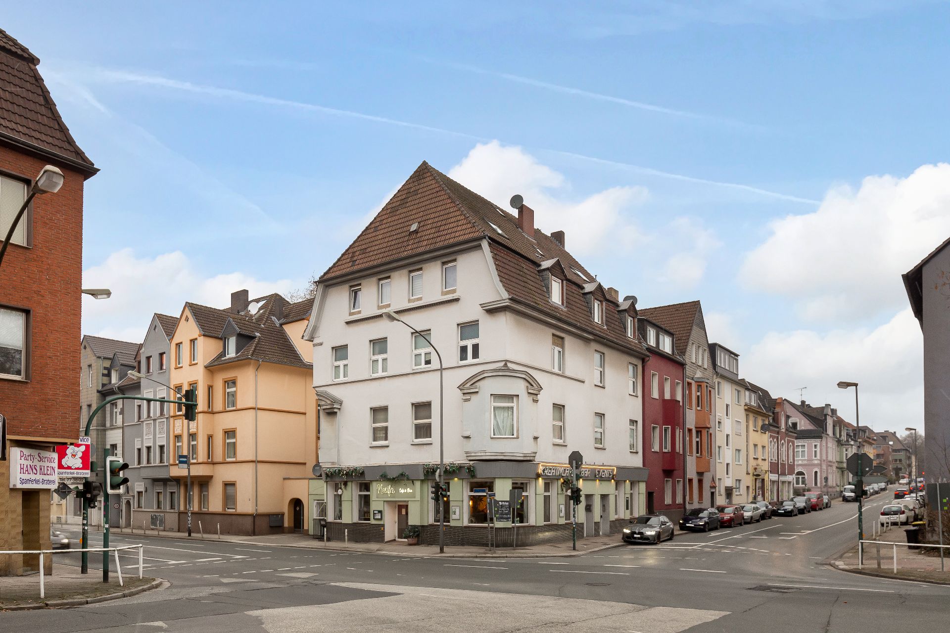 Mehrstöckiges Wohngebäude an einer Straßenecke mit geparkten Autos und weiteren Häusern im Hintergrund.