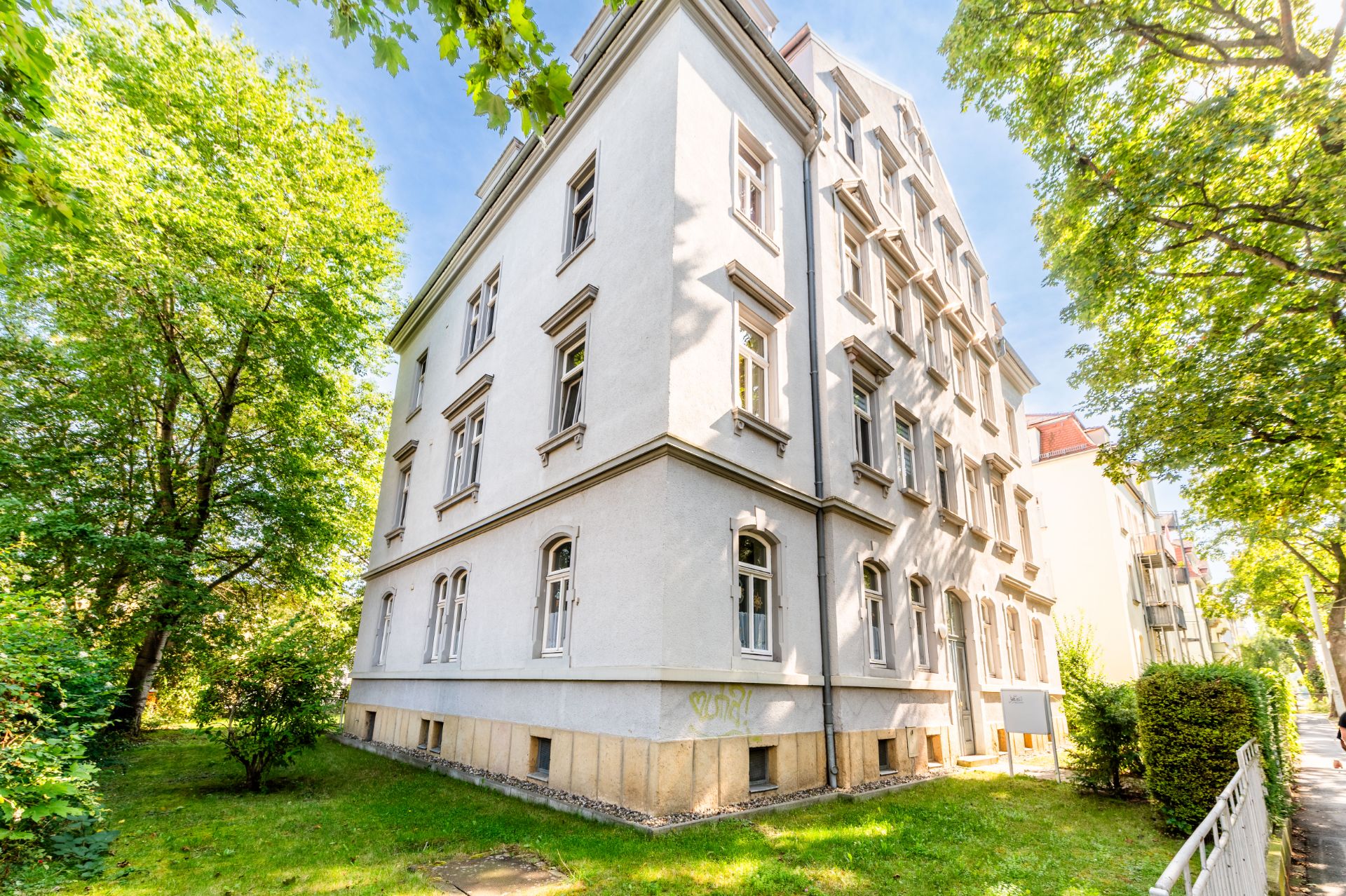 Weißes mehrstöckiges Wohngebäude mit vielen Fenstern, umgeben von Bäumen und Grünfläche.