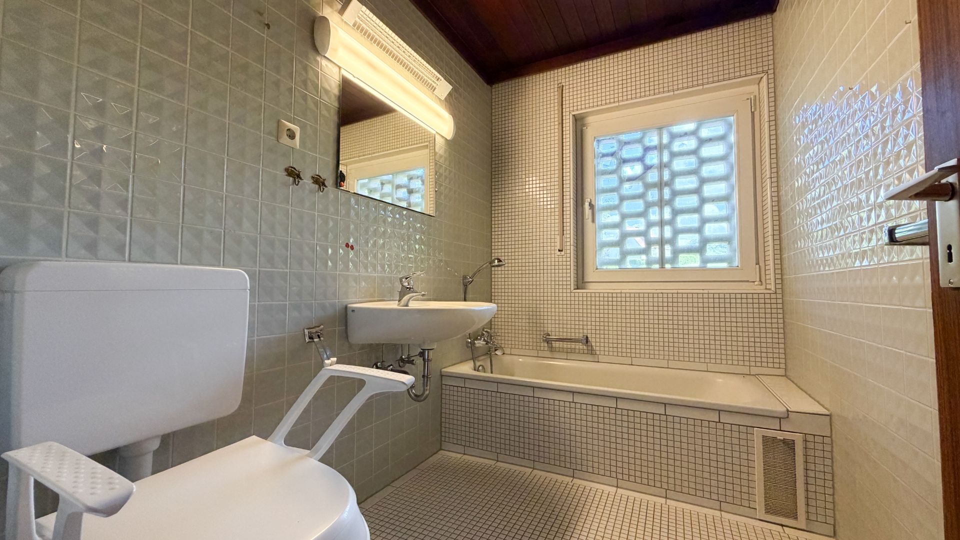 Badezimmer mit Toilette, Waschbecken, Badewanne und Fenster, weiße Fliesen an Wand und Boden.