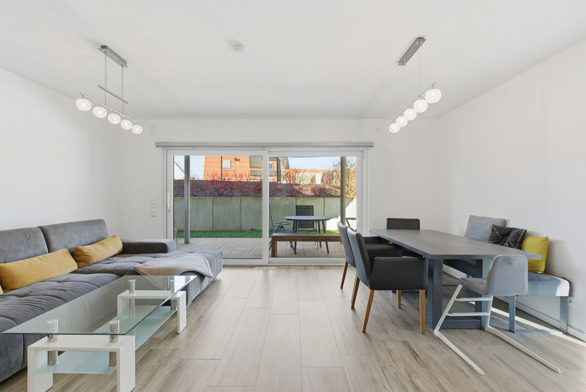 Modernes Wohnzimmer mit grauem Sofa, Esstisch, Glastischen und Zugang zur Terrasse mit Garten.