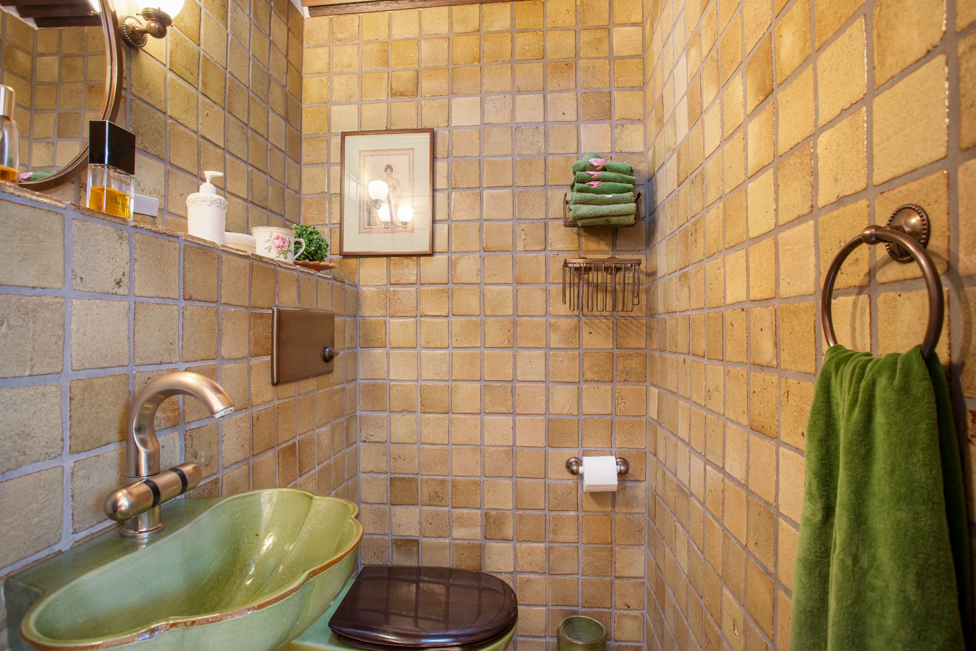 Kleines Badezimmer mit braunen Fliesen, grünem Waschbecken, Toilette und grünen Handtüchern.