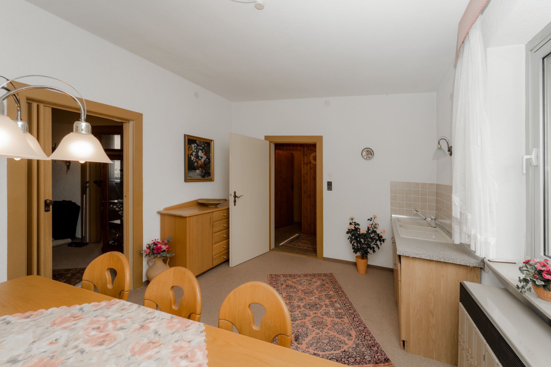Esszimmer mit Holztisch, Stühlen, Kommode, Teppich und Fenster mit Gardinen.