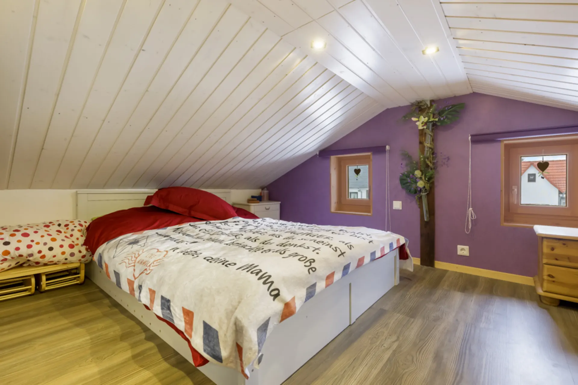 Gemütliches Schlafzimmer mit Dachschräge, lila Wand, Doppelbett und zwei Fenstern.
