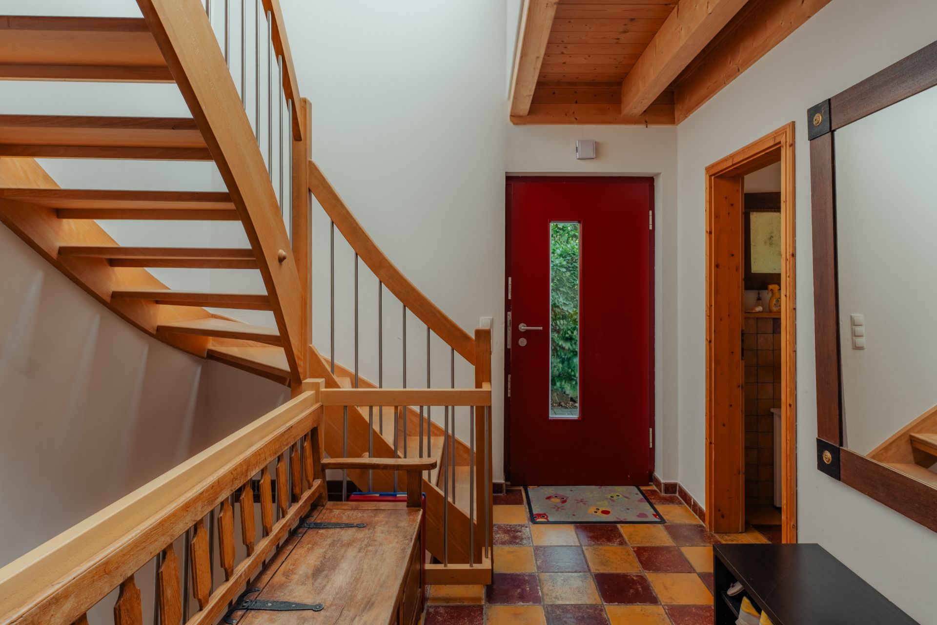 Flur mit roter Tür, Holztreppe, Spiegel und Sitzbank, Fliesenboden in warmen Farben.