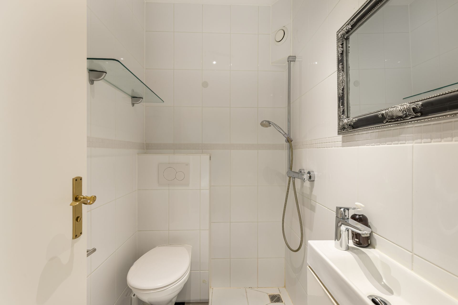 Kleines weiß gefliestes Badezimmer mit Toilette, Dusche, Waschbecken und Spiegel.