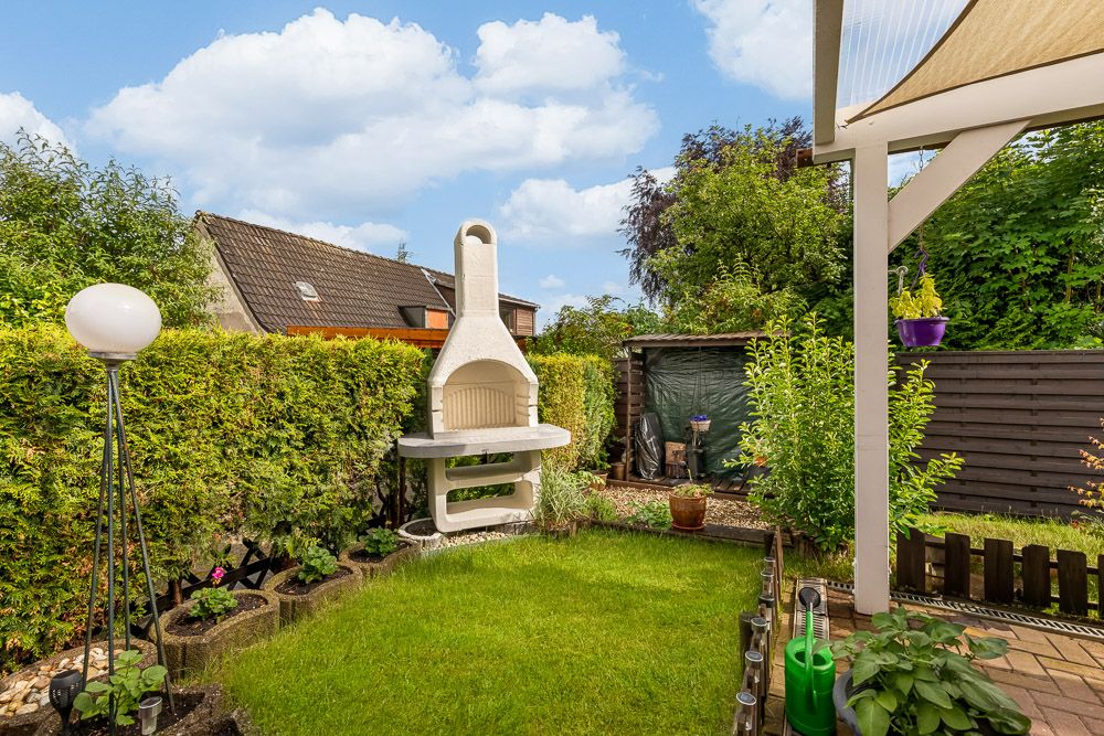 Kleiner Garten mit Rasen, weißem Grill, Hecke und Gartenhaus bei Tageslicht.