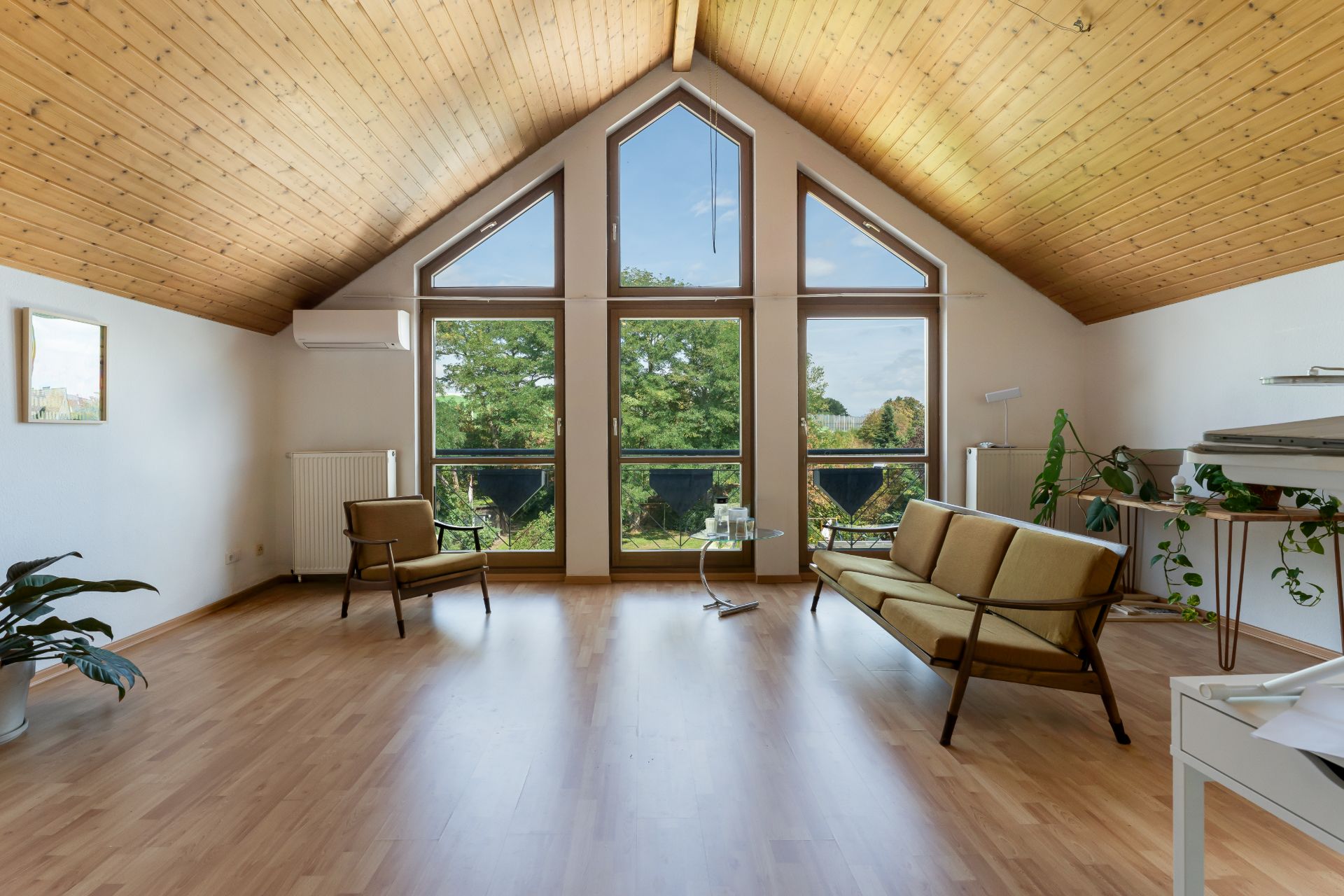 Helles Wohnzimmer mit Holzdecke, großen Fenstern, zwei Sesseln, Sofa und Pflanzen.