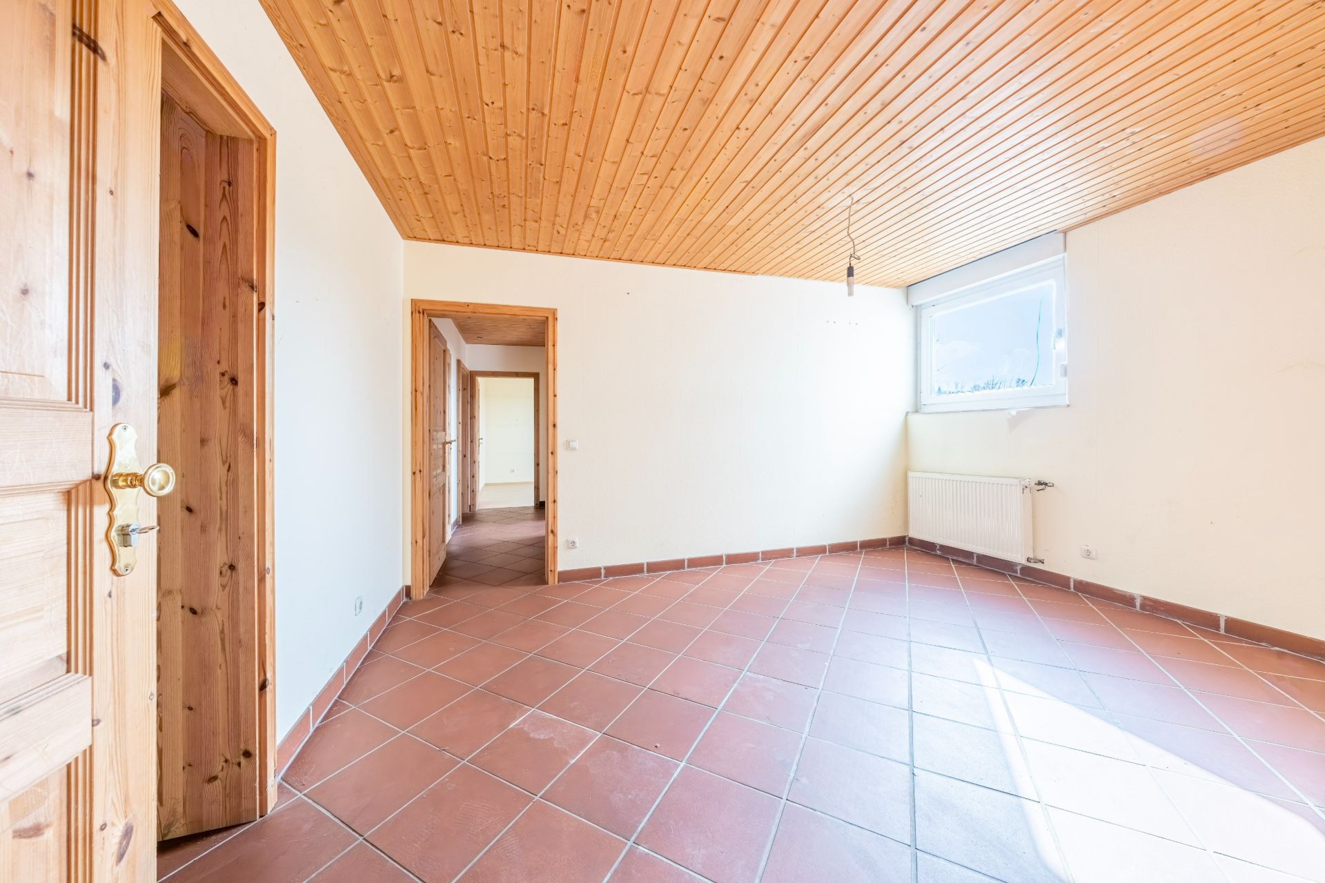 Leerer Raum mit Fliesenboden, Holzdecke, Türöffnungen und großem Fenster mit Heizung darunter.
