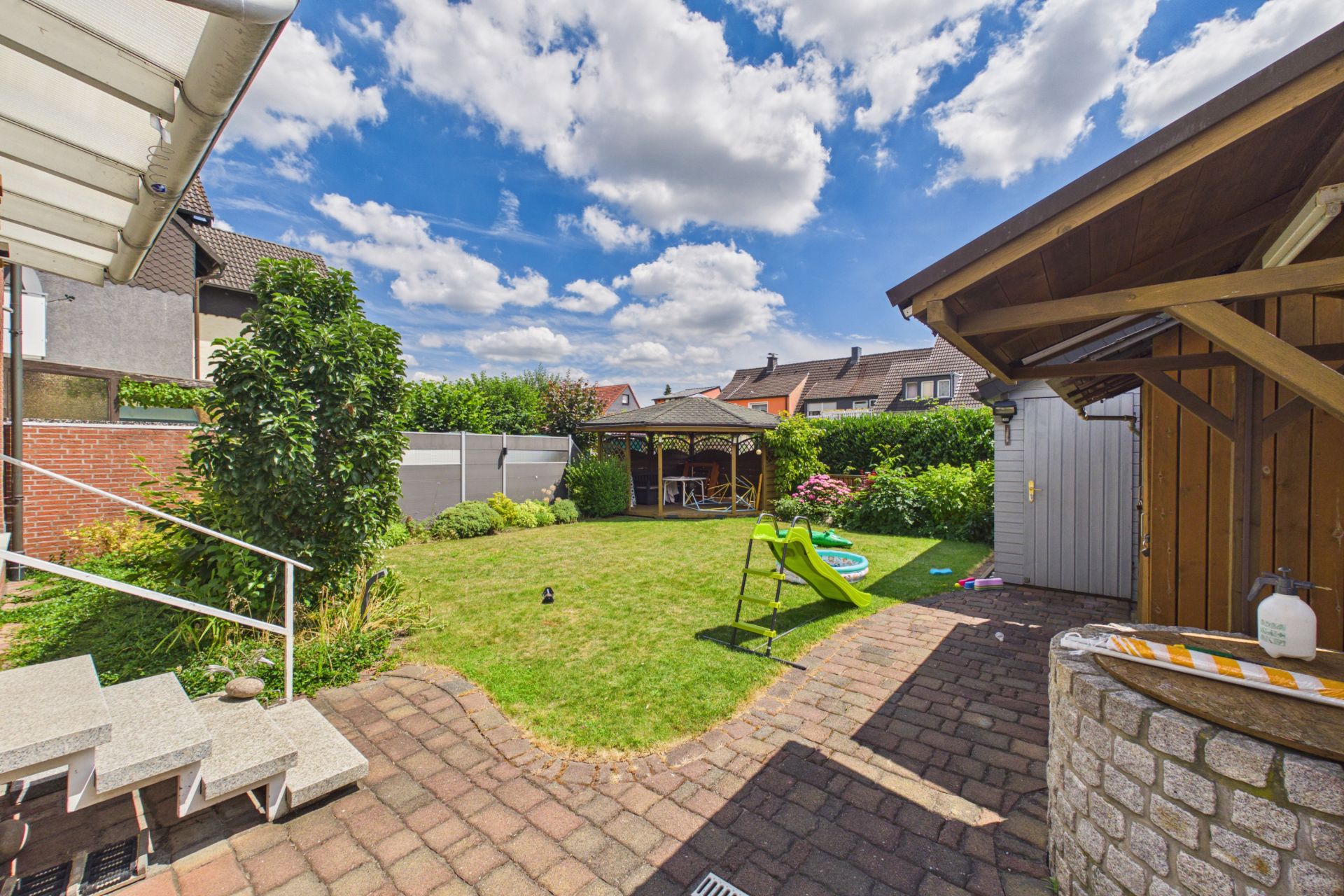 Garten mit Rasen, Rutsche, kleinem Pavillon, Schuppen und Terrasse bei sonnigem Wetter.