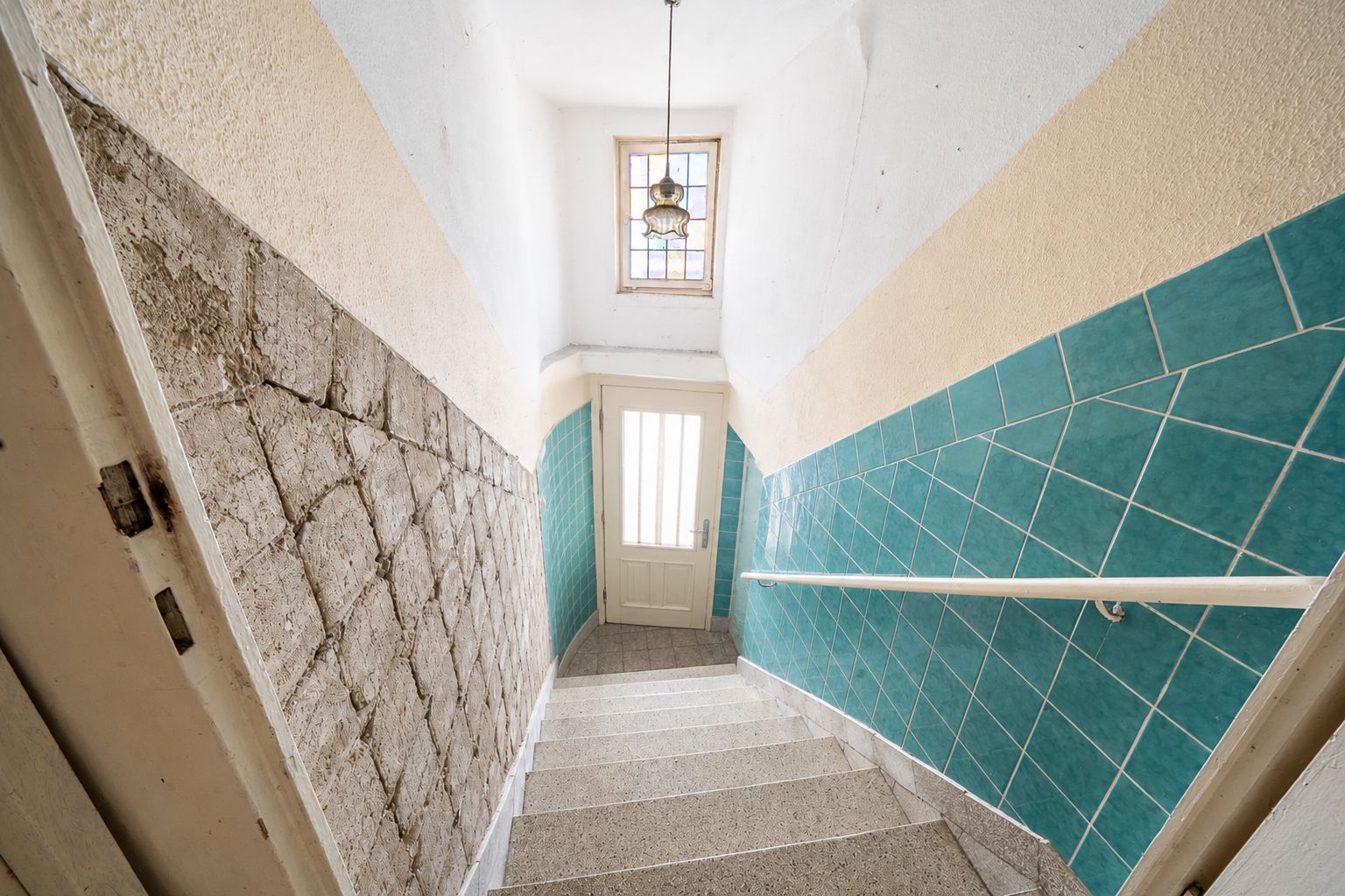 Treppe mit grün gefliester Wand und Fenster am unteren Ende, Tür mit Glaseinsatz sichtbar.