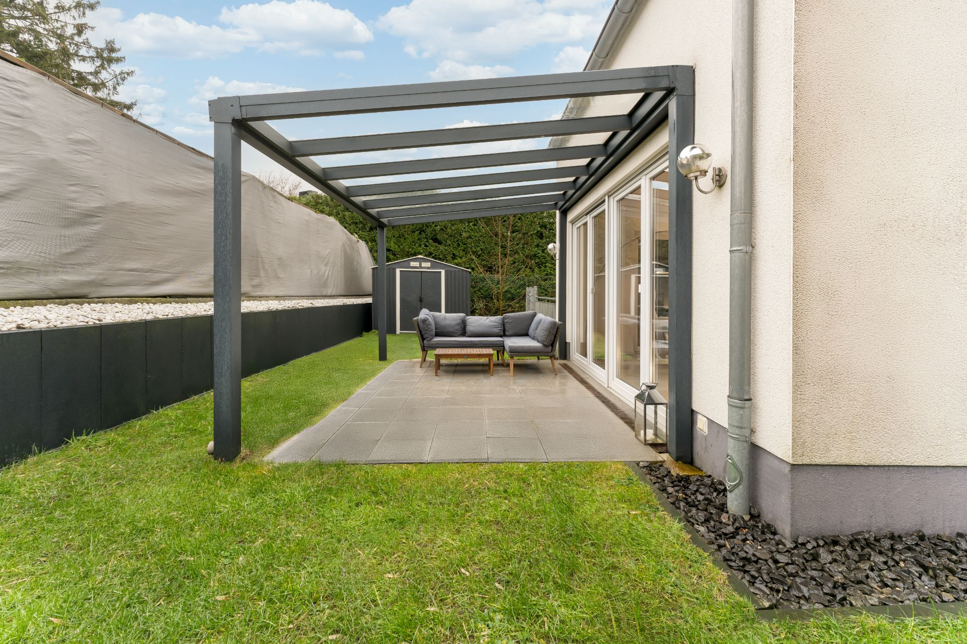 Überdachte Terrasse mit Sofa und Tisch im Garten neben einem modernen Haus.