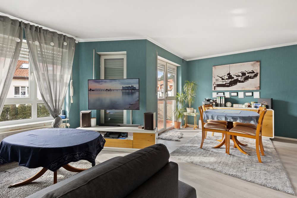 Helles Wohnzimmer mit zwei Esstischen, Fernseher, großen Fenstern und grünblauen Wänden.