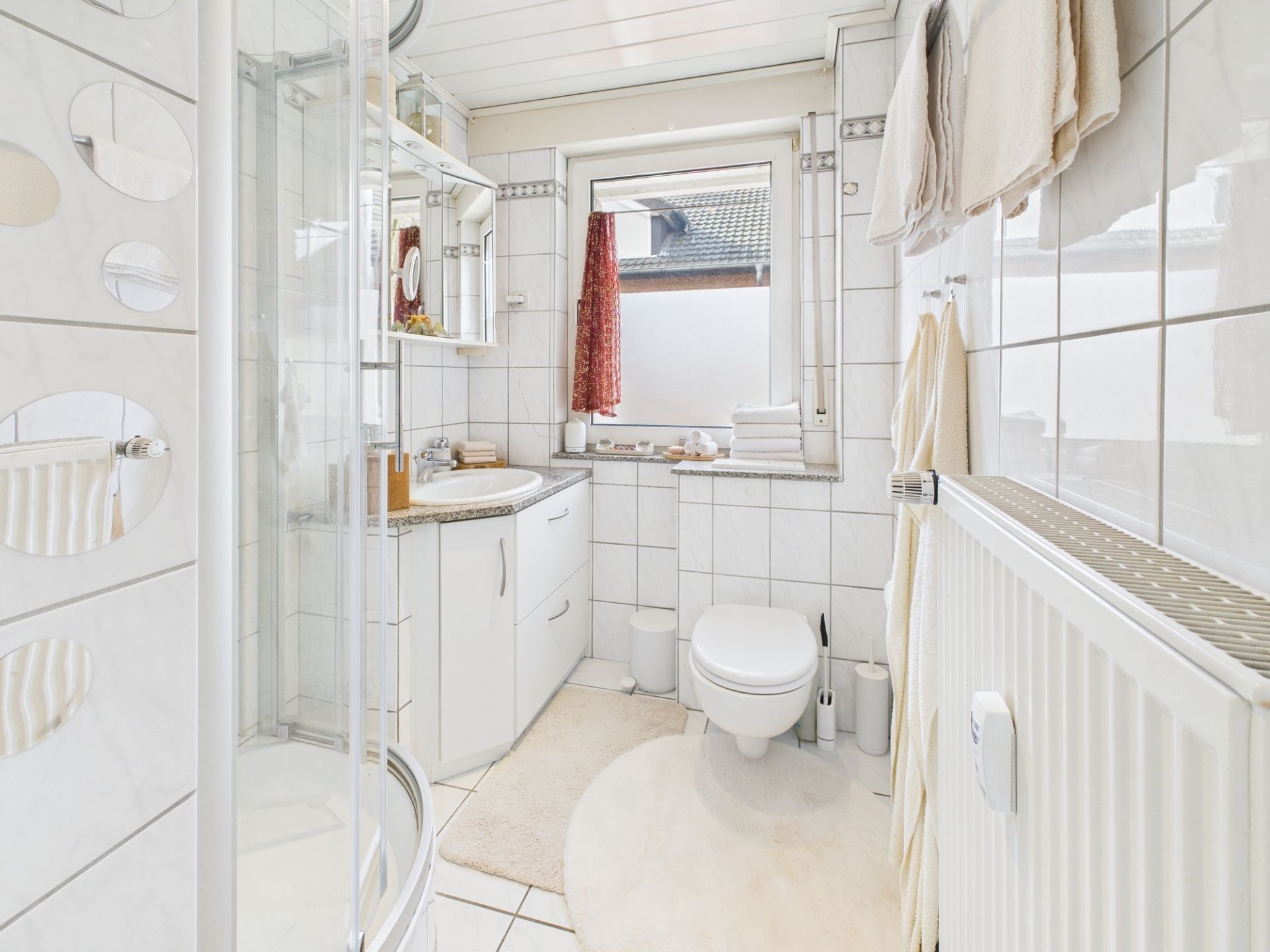Helles Badezimmer mit Dusche, Waschbecken, WC, Fenster und Handtüchern auf weiß gefliesten Wänden.