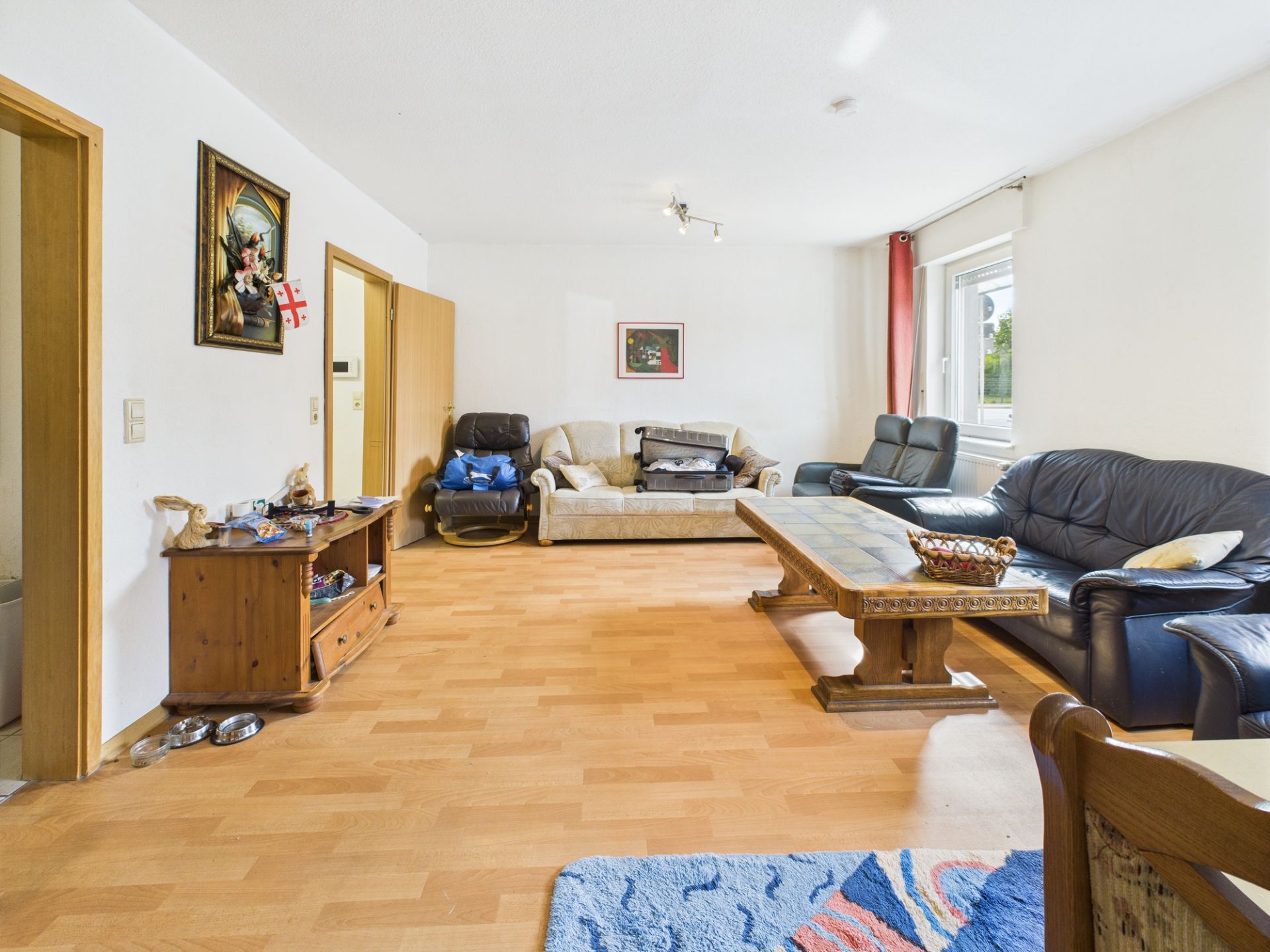 Helles Wohnzimmer mit drei Sofas, Holztisch, Kommode und großem Fenster.