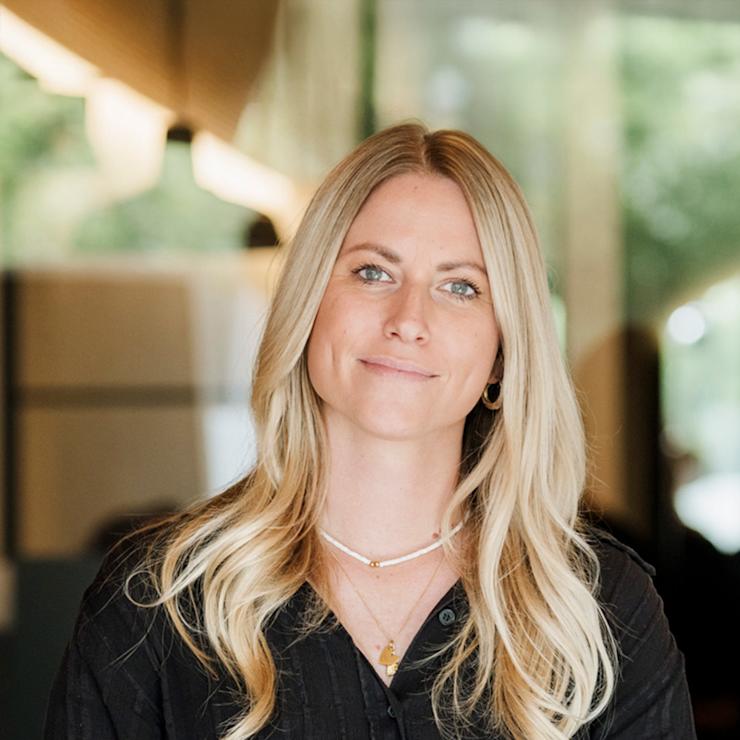 Junge Frau mit langen blonden Haaren trägt einen dunklen Blazer und steht draußen.