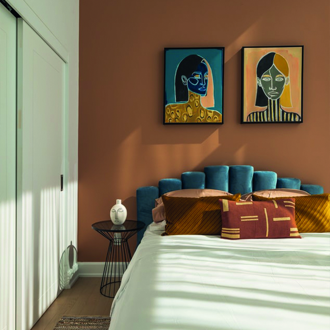 Schlafzimmer mit blauem Bett, Kissen, Nachttisch und zwei abstrakten Porträts an brauner Wand.