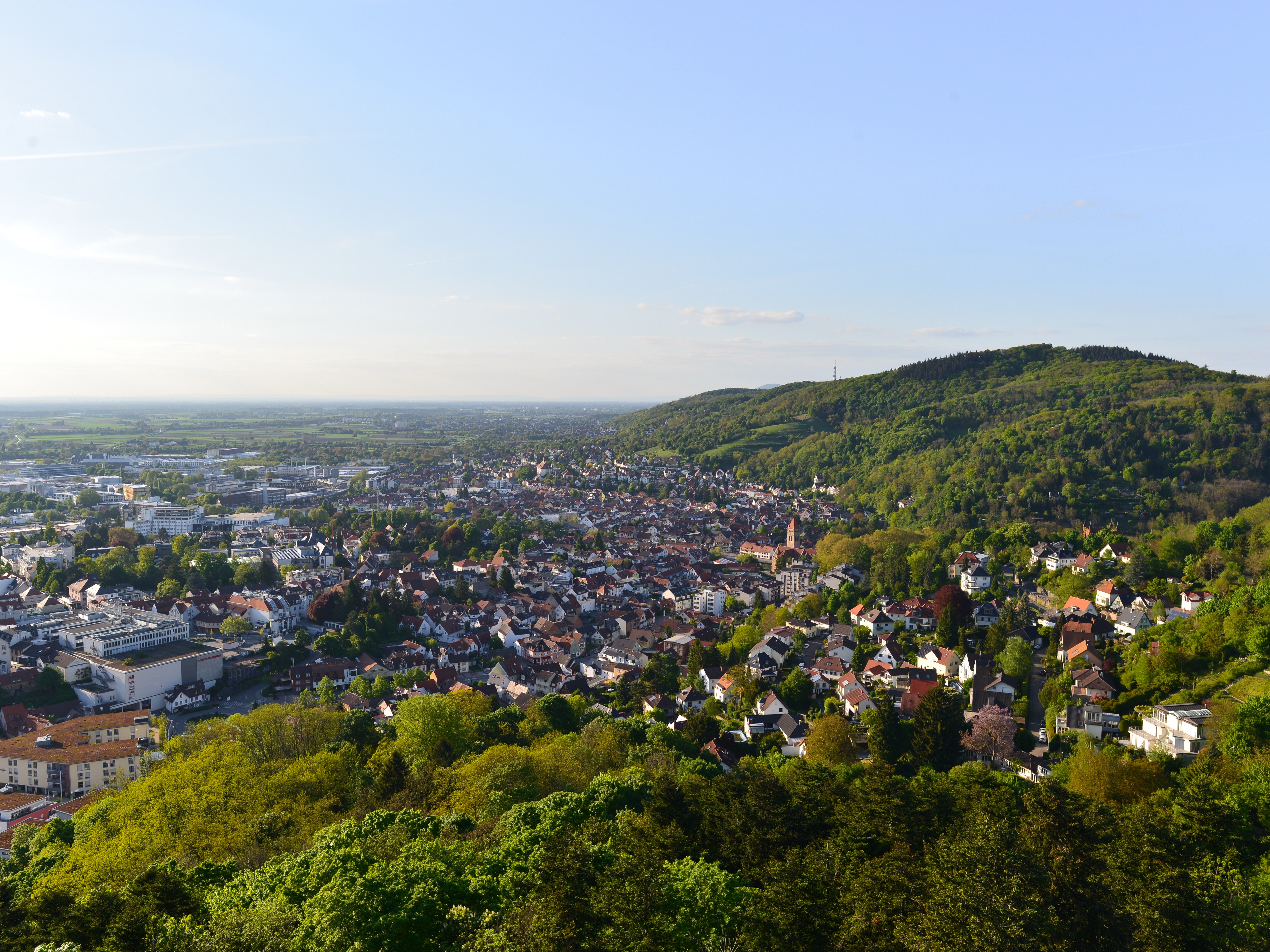 Blick über eine Stadt mit vielen Häusern, Grünflächen und bewaldeten Hügeln im Hintergrund.