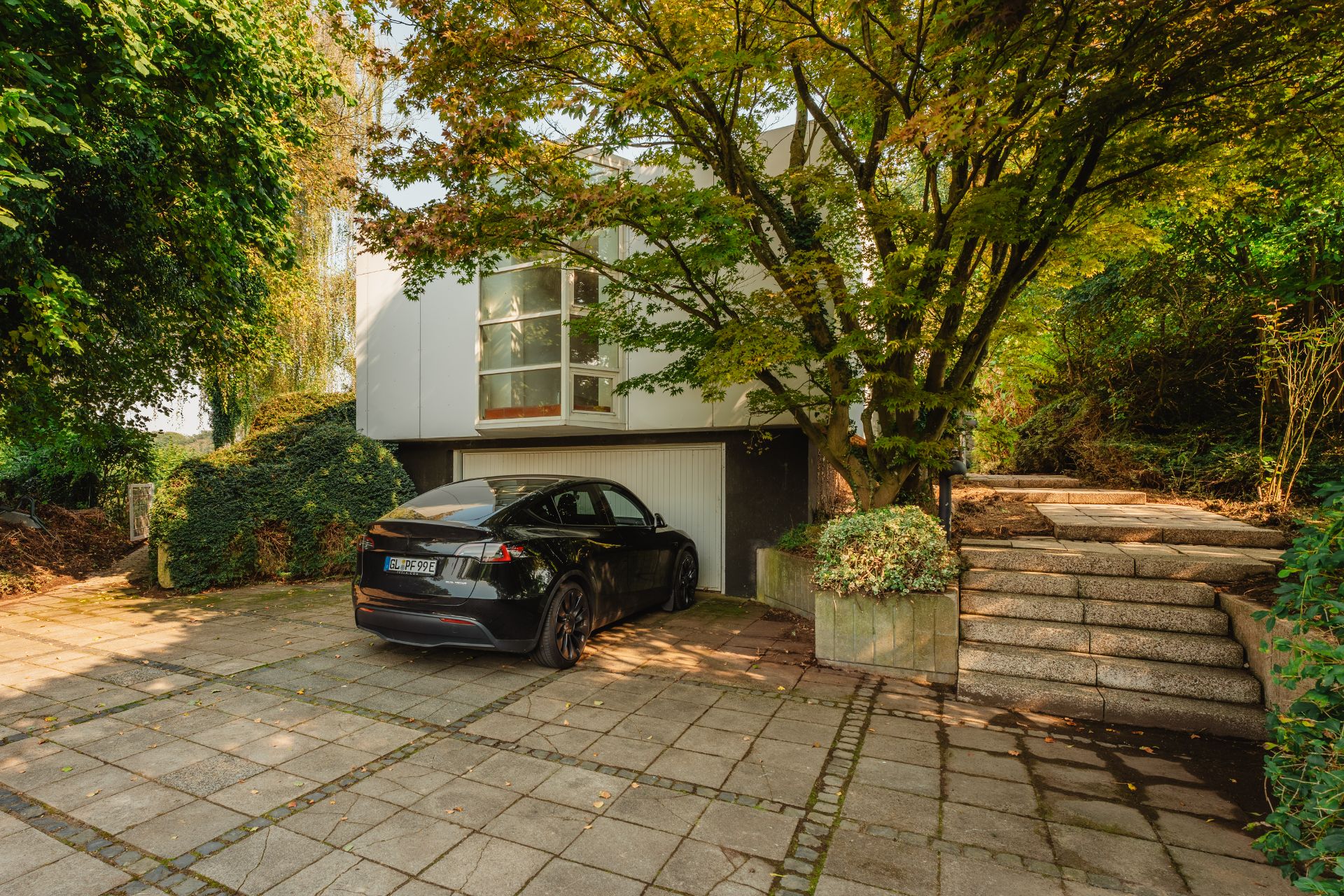 Ein schwarzes Auto parkt vor einer modernen Wohnhausgarage, daneben führen Stufen durch grüne Bäume.