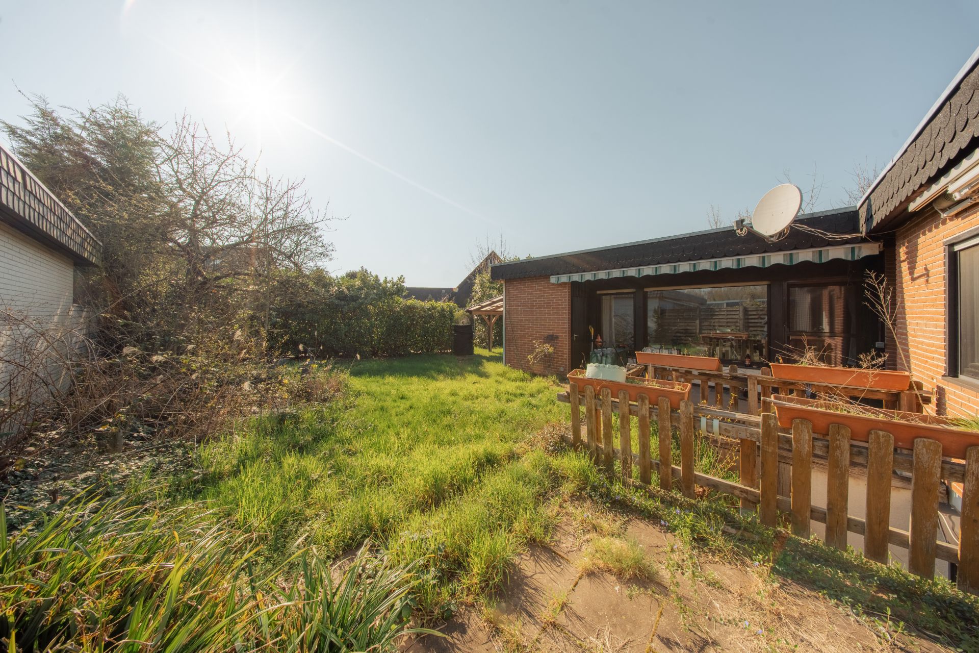 Garten mit Terrasse und Holzzaun vor einem Backsteinhaus bei sonnigem Wetter.
