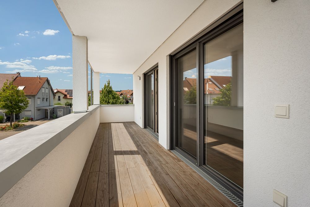 Großer Balkon mit Holzboden, Glas-Schiebetüren und Blick auf Wohnhäuser.