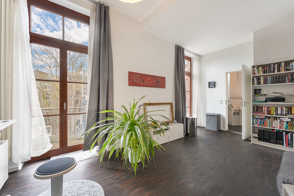 Helles Wohnzimmer mit großen Fenstern, Pflanze, Bücherregal und dunklem Holzboden.