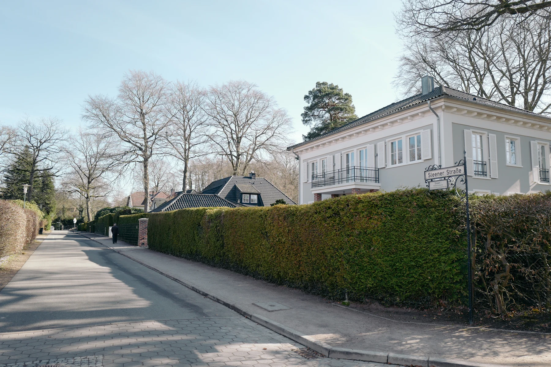 Wohnhaus mit Balkon und weißen Fensterläden an einer ruhigen, baumbestandenen Straße.