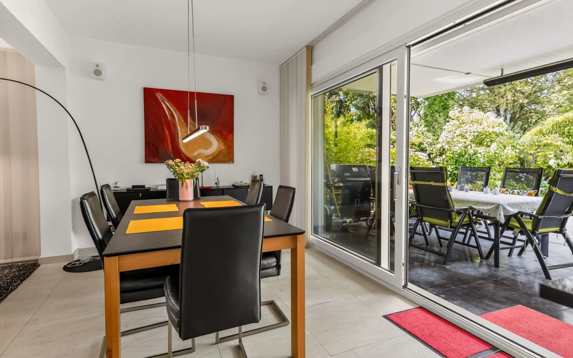 Esszimmer mit schwarzem Tisch, sechs Stühlen, rotem Bild und Zugang zur Terrasse mit Sitzgruppe.