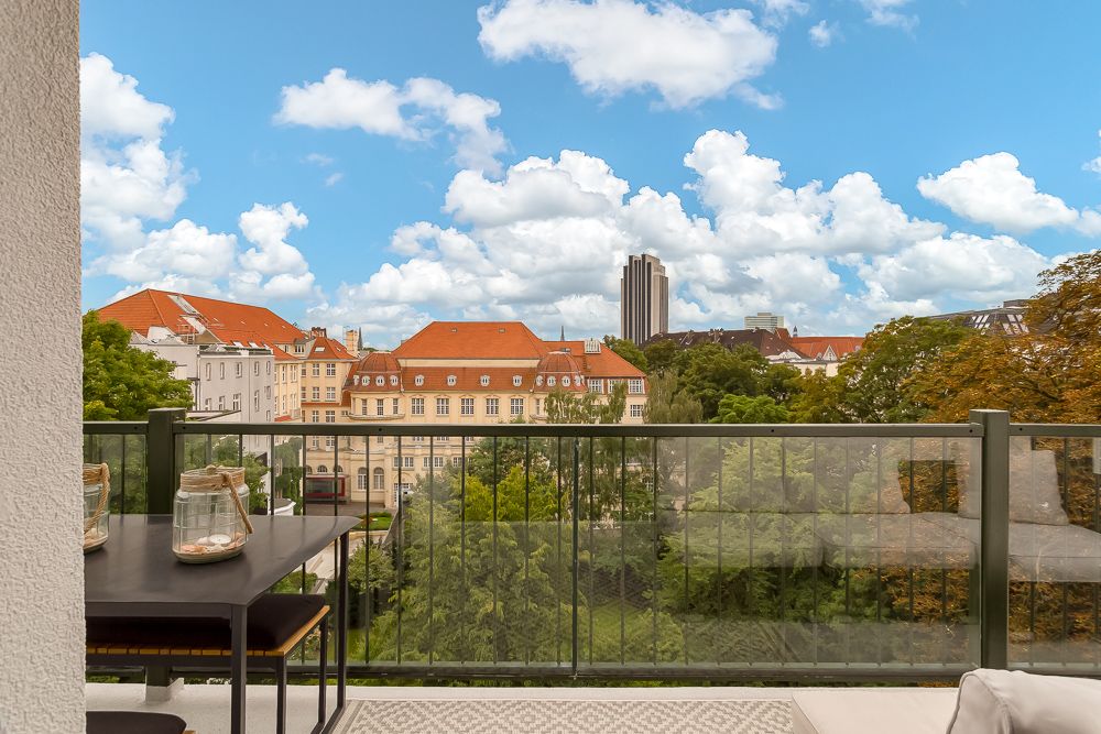 Balkon mit Glasgeländer, Tisch und Stühlen, Blick auf Bäume und Gebäude mit roten Dächern.