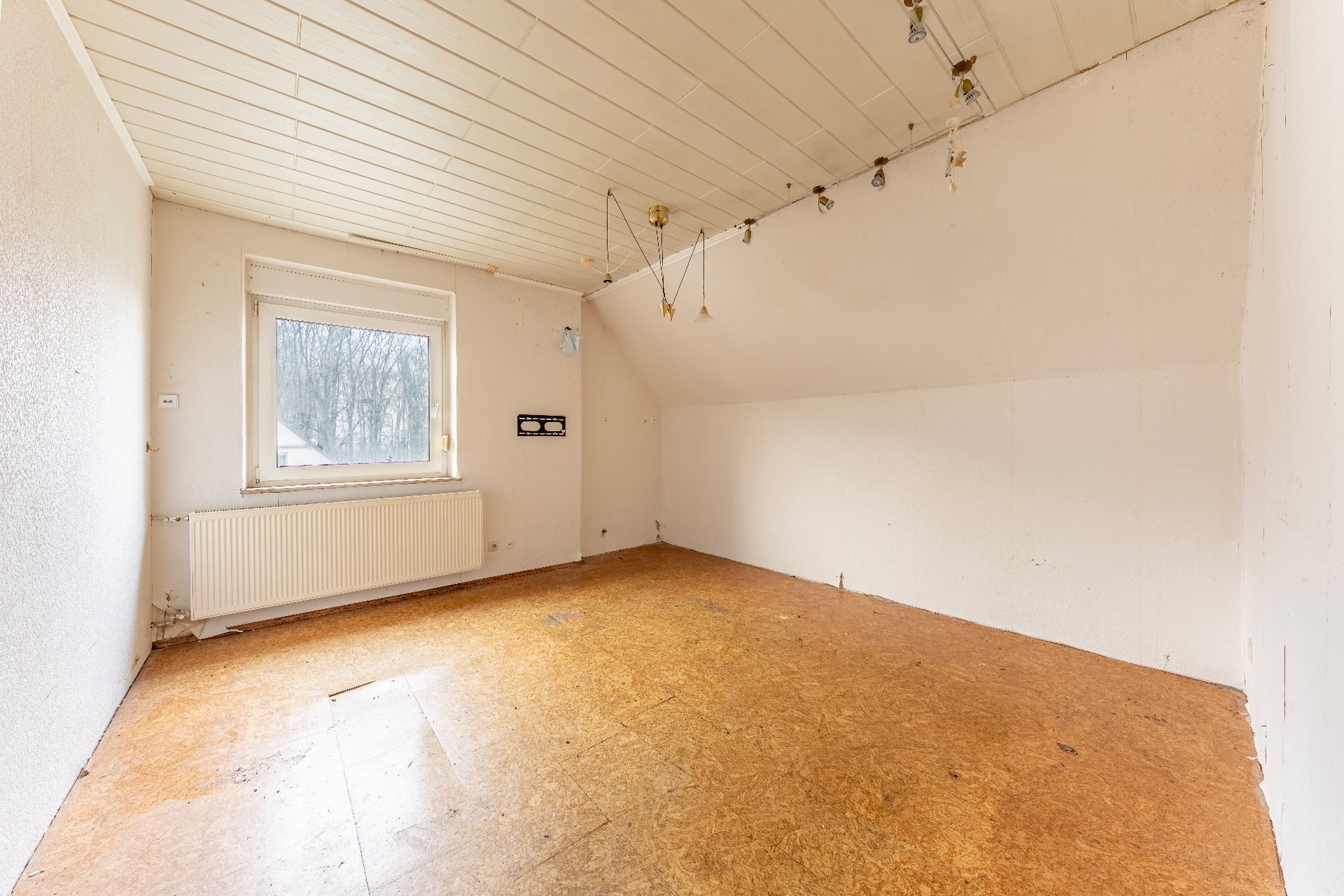 Leeres Zimmer mit schräger Decke, Fenster, Heizung und braunem Boden.