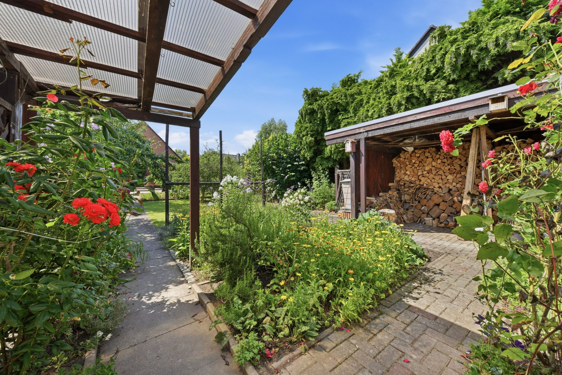 Garten mit Blumen, überdachtem Weg und Holzunterstand mit gestapeltem Brennholz.