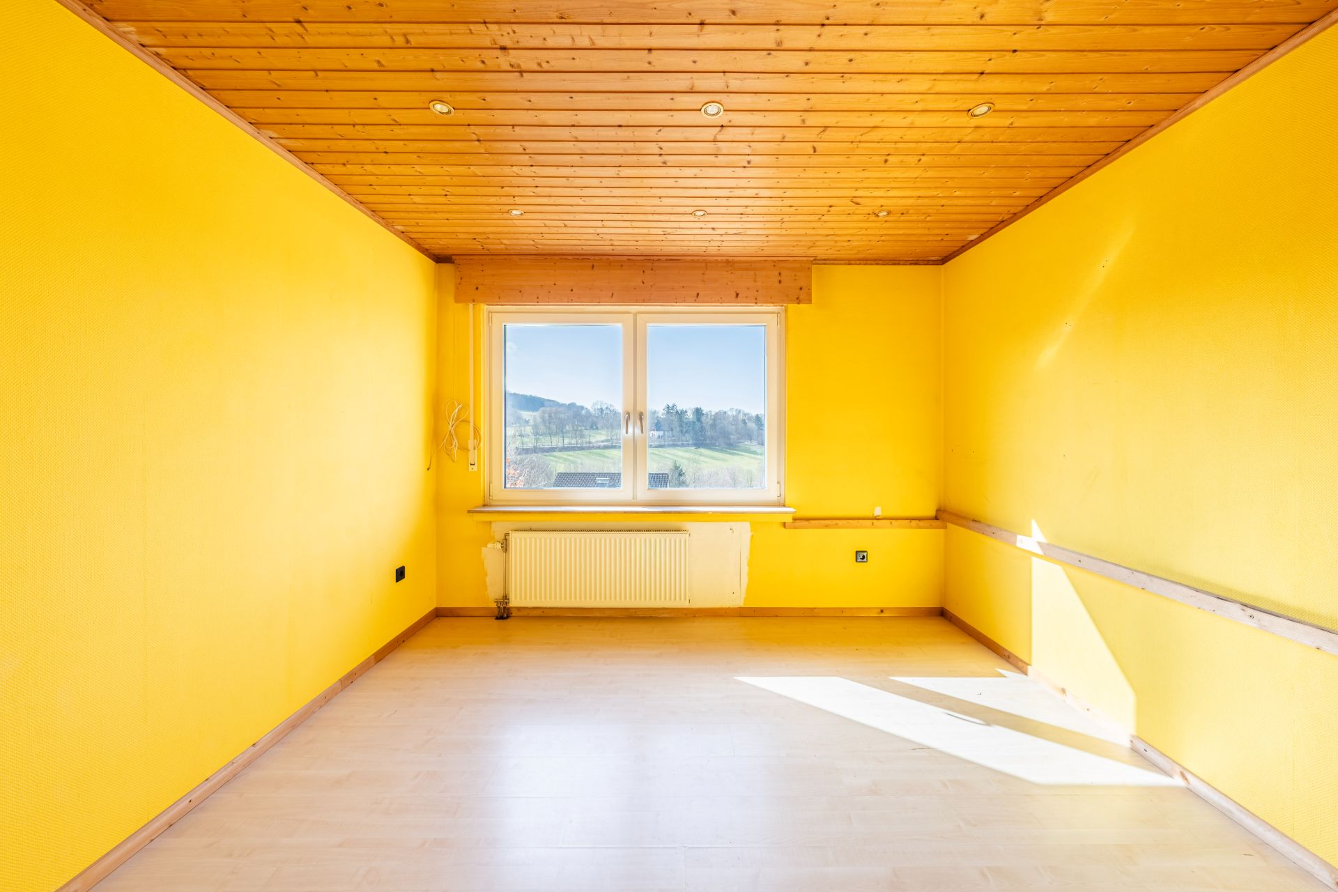 Leerer Raum mit gelben Wänden, Holzboden und Decke, großem Fenster und Heizkörper.