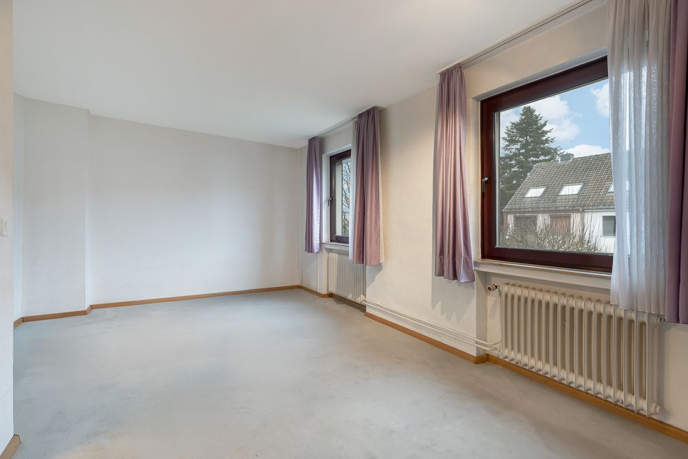Leeres Zimmer mit zwei Fenstern, lila Vorhängen und Heizkörper unter den Fenstern.