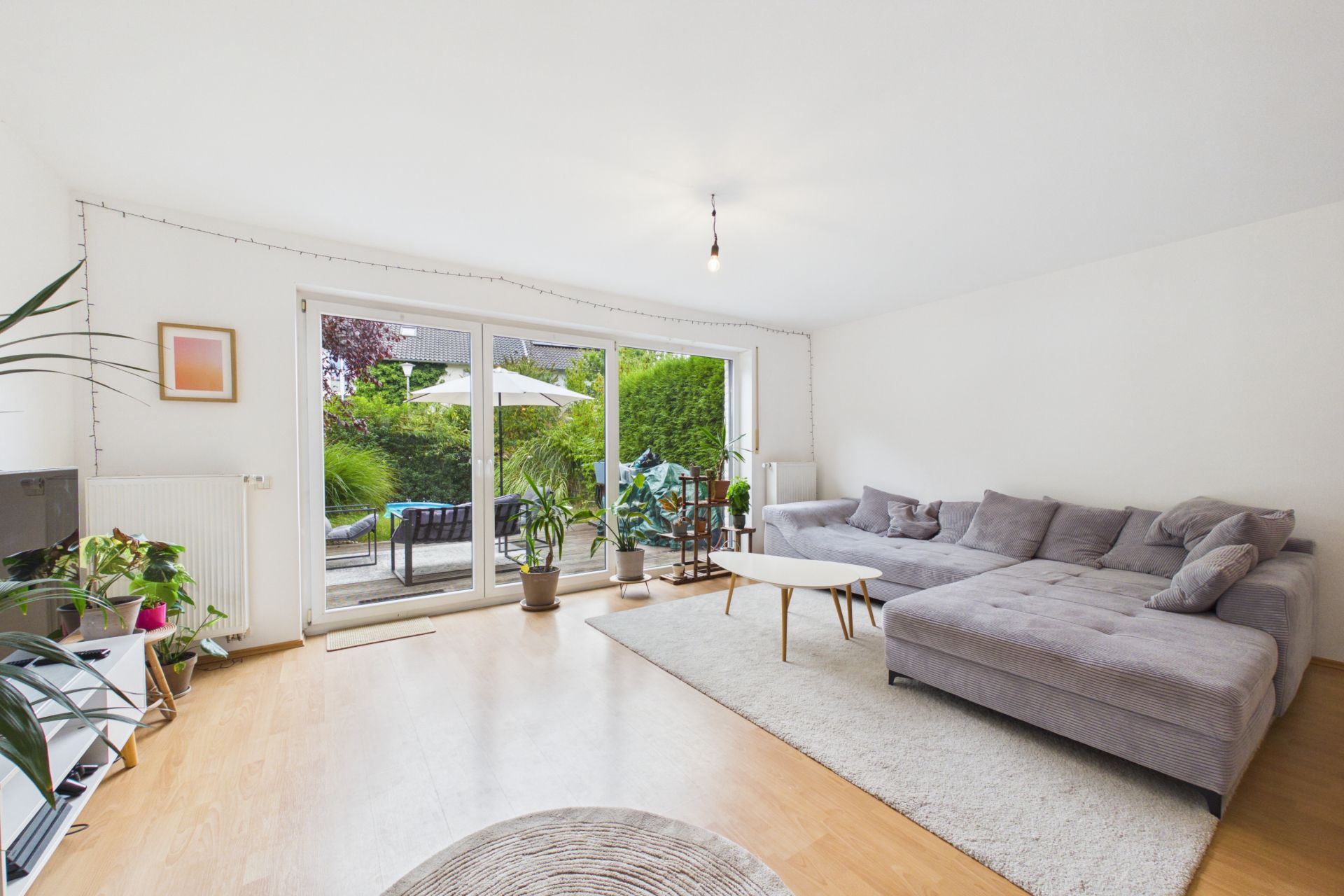 Helles Wohnzimmer mit grauem Ecksofa, Couchtisch, Pflanzen und Zugang zur Terrasse mit Garten.