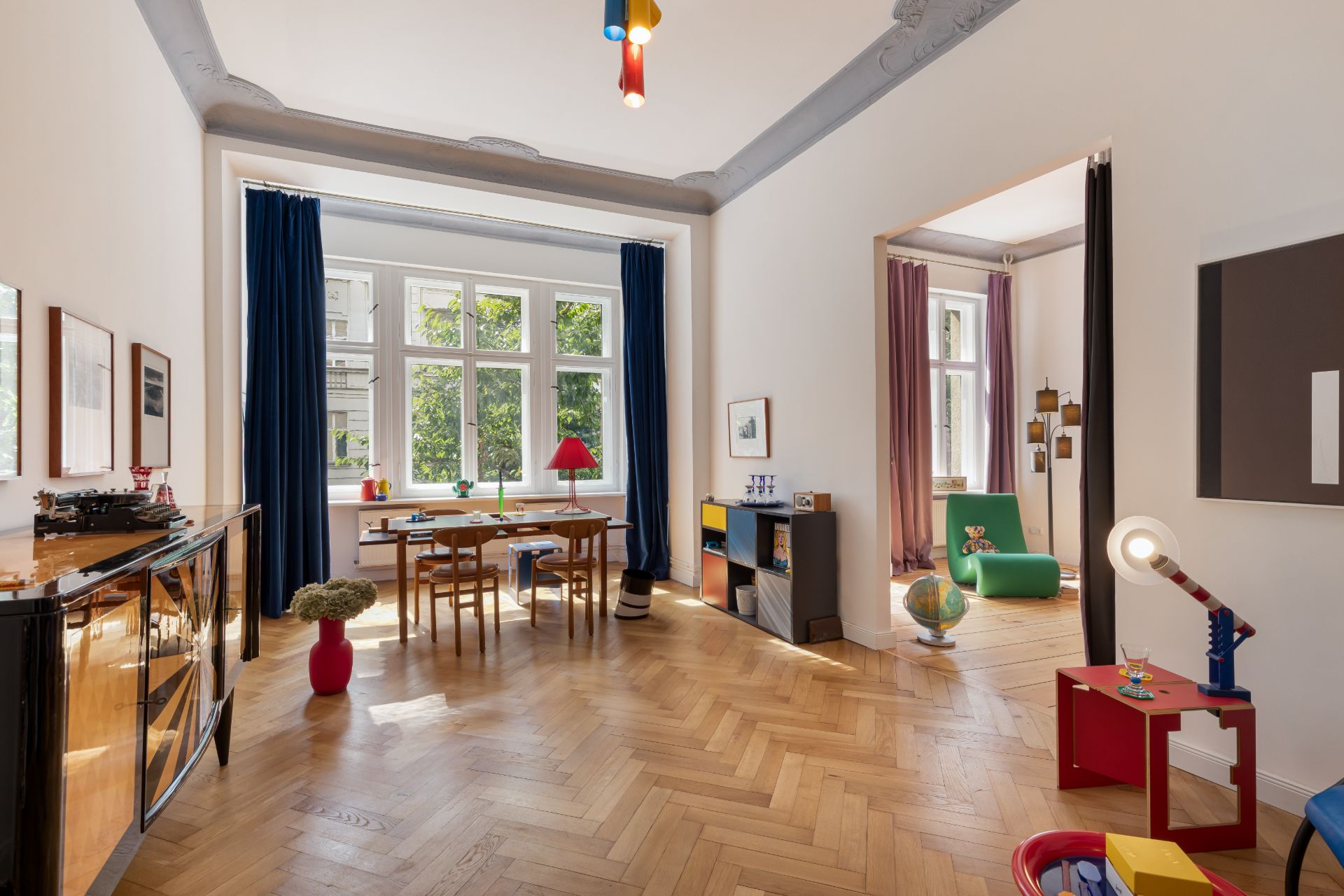 Helles Wohnzimmer mit Holzboden, großen Fenstern, Kindermöbeln und bunten Vorhängen.