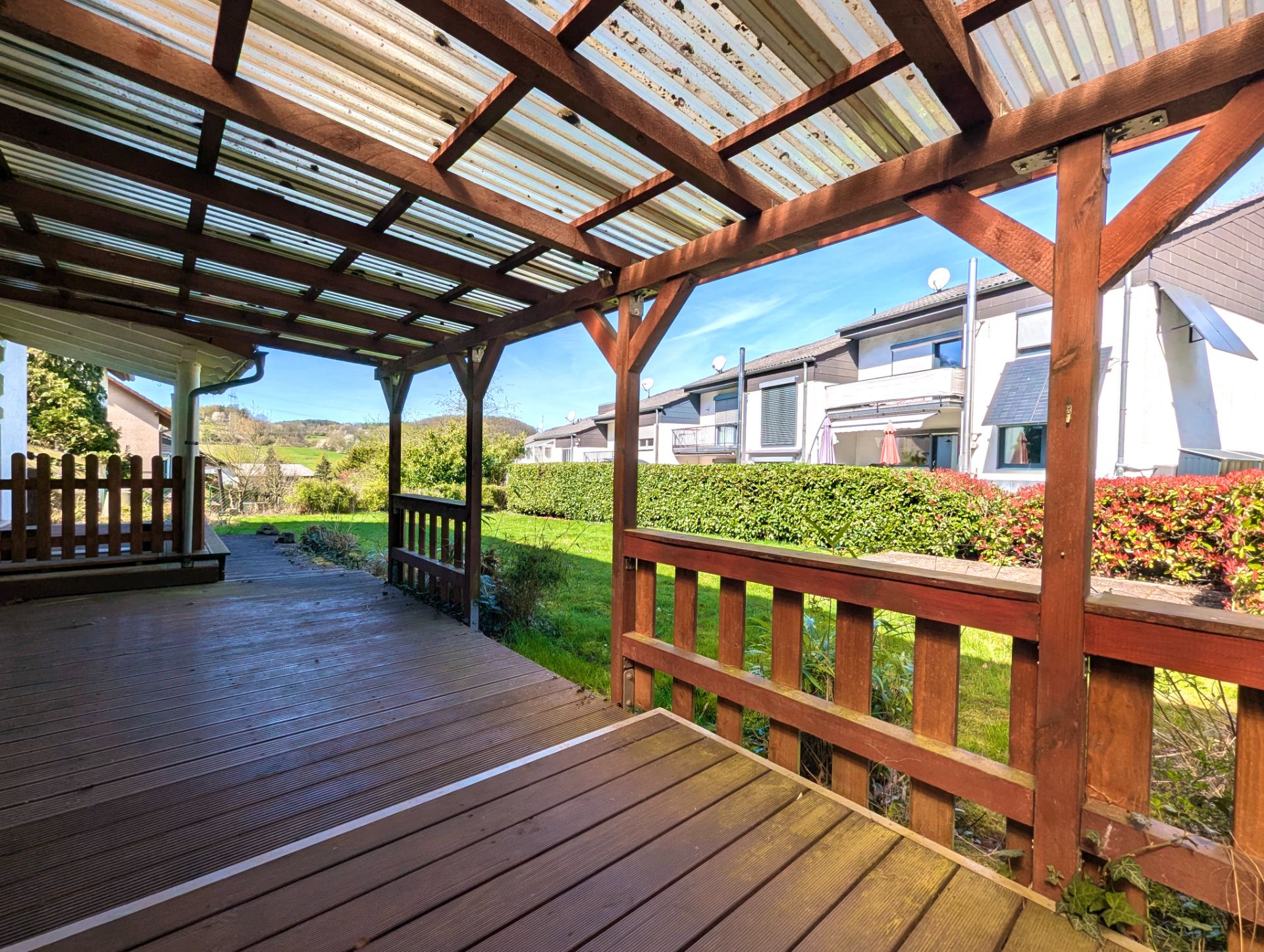 Überdachte Holzterrasse mit Blick auf Garten und benachbarte Häuser.