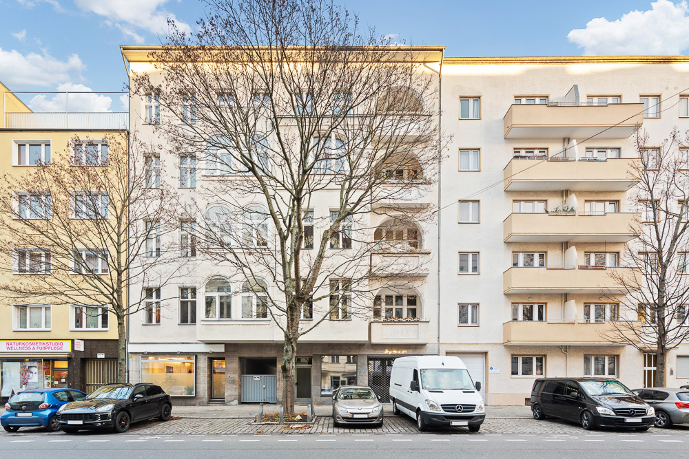 Mehrstöckiges Wohnhaus mit Balkonen und geparkten Autos davor, Bäume ohne Blätter am Straßenrand.