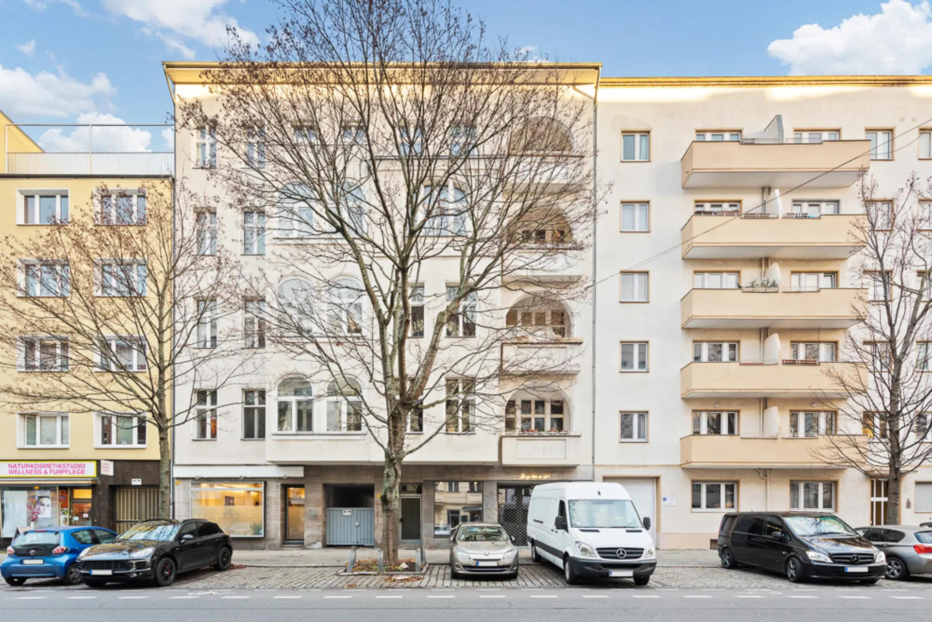 Mehrstöckiges Wohnhaus mit Balkonen und geparkten Autos davor, Bäume ohne Blätter am Straßenrand.