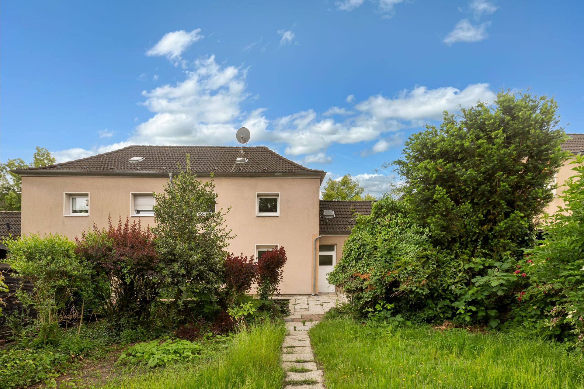 Rückansicht eines Hauses mit Garten und Weg, umgeben von Büschen und Bäumen bei blauem Himmel.